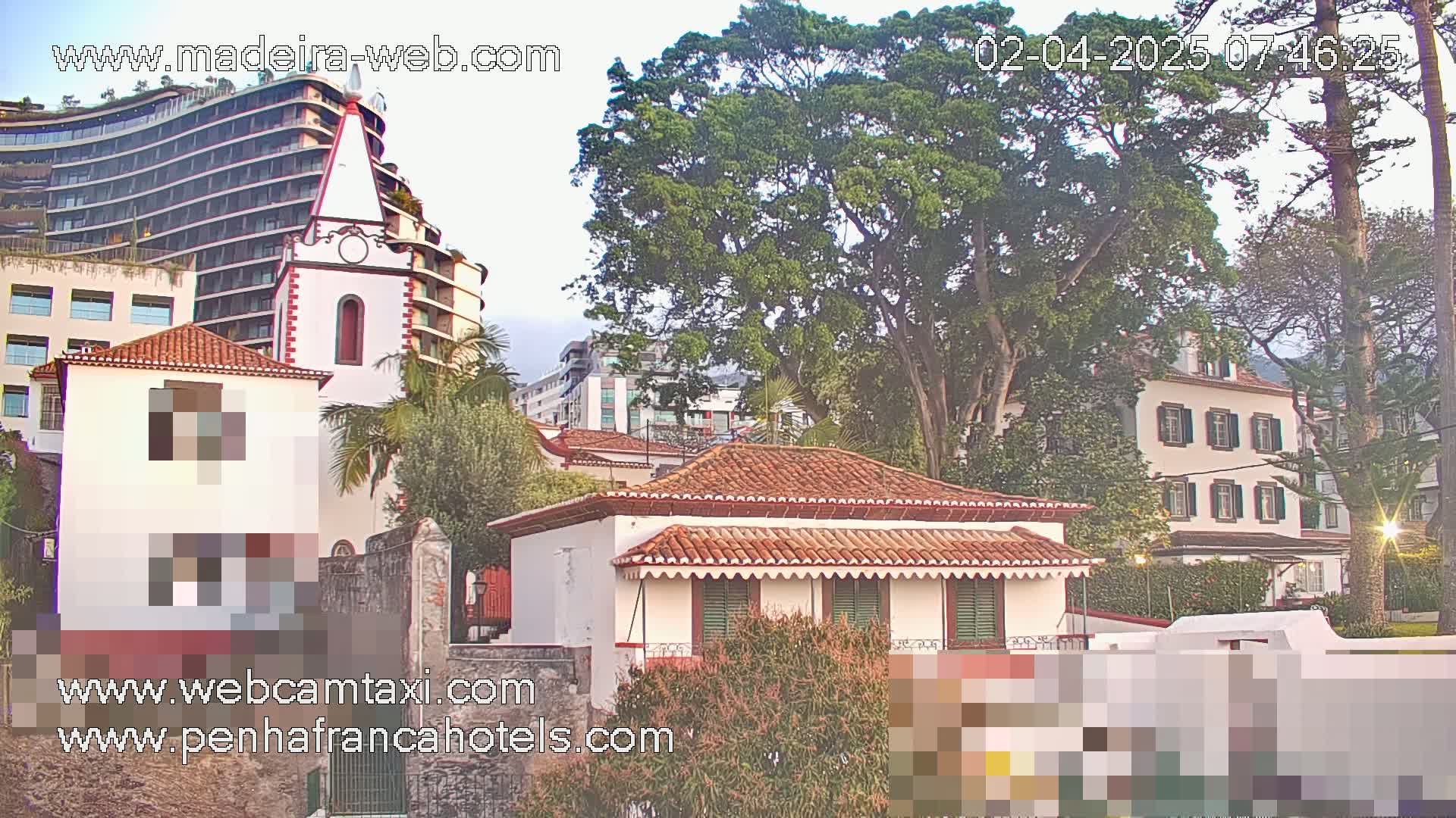 A small, white house with a red tile roof sits among taller buildings and lush green trees under a partly cloudy sky.