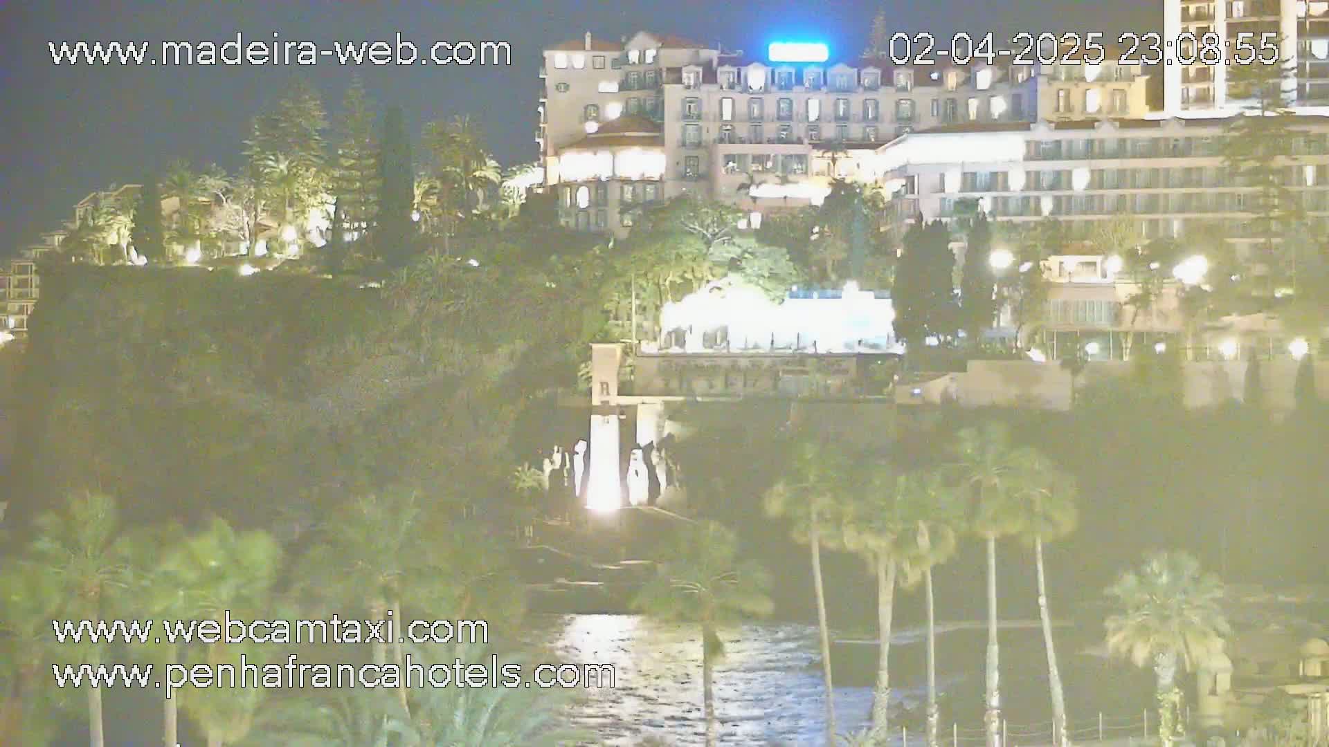 A nighttime view of a waterfront hotel complex, illuminated, with lush vegetation and palm trees surrounding a calm body of water.