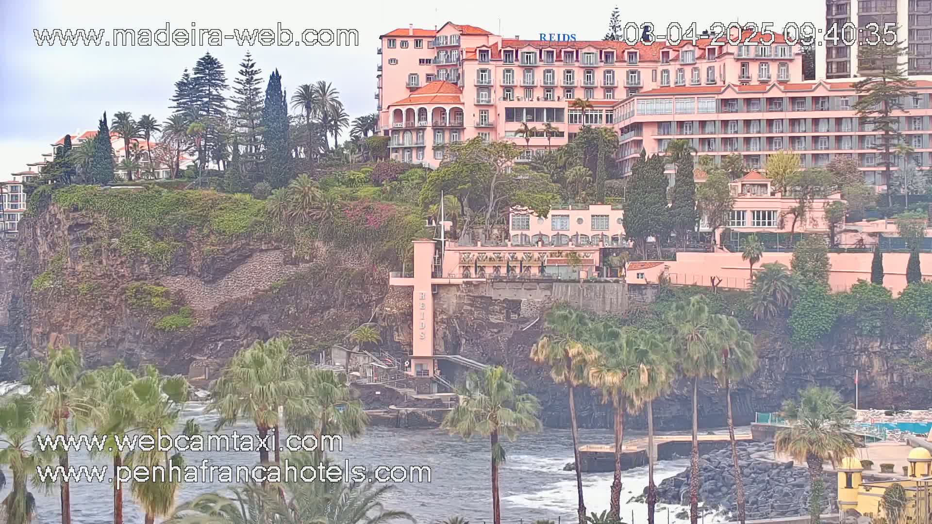 A large pink hotel sits atop a rocky cliff overlooking a choppy ocean, surrounded by lush green vegetation and palm trees under an overcast sky.