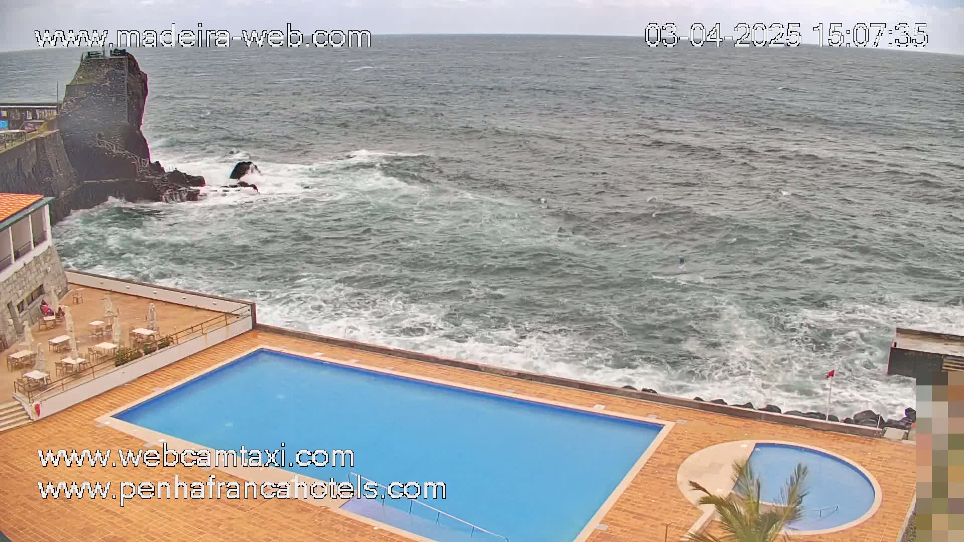 A hotel pool and patio area overlook a rocky coastline with choppy ocean waves under an overcast sky.