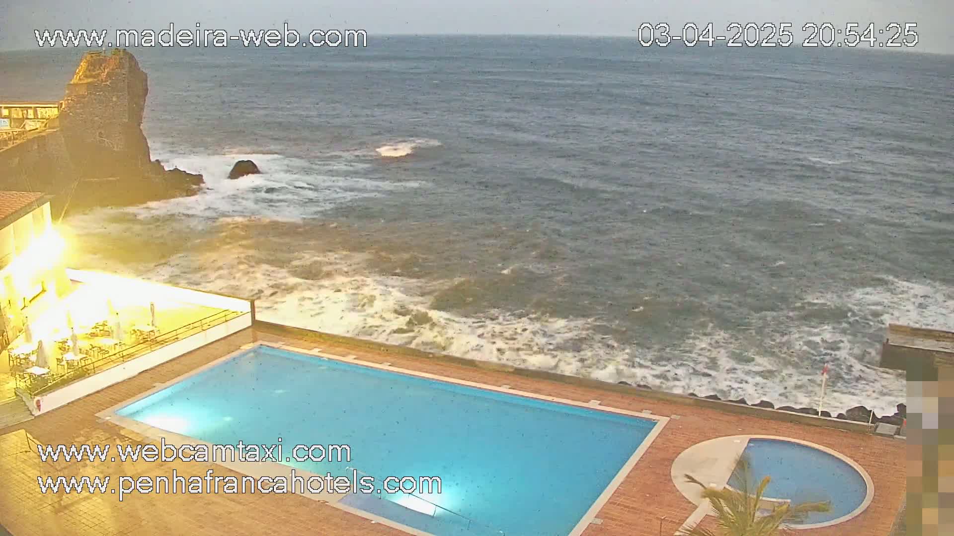 An outdoor swimming pool and smaller circular pool are situated next to a rocky coastline with moderately rough ocean waves under an overcast sky.