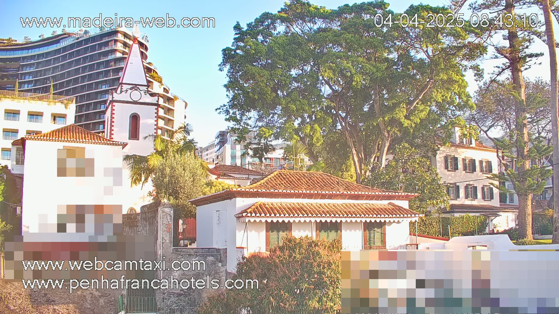 A sunny day reveals a cluster of buildings, including a multi-story hotel, a church-like structure, and smaller houses with terracotta roofs, all nestled amongst lush green trees.