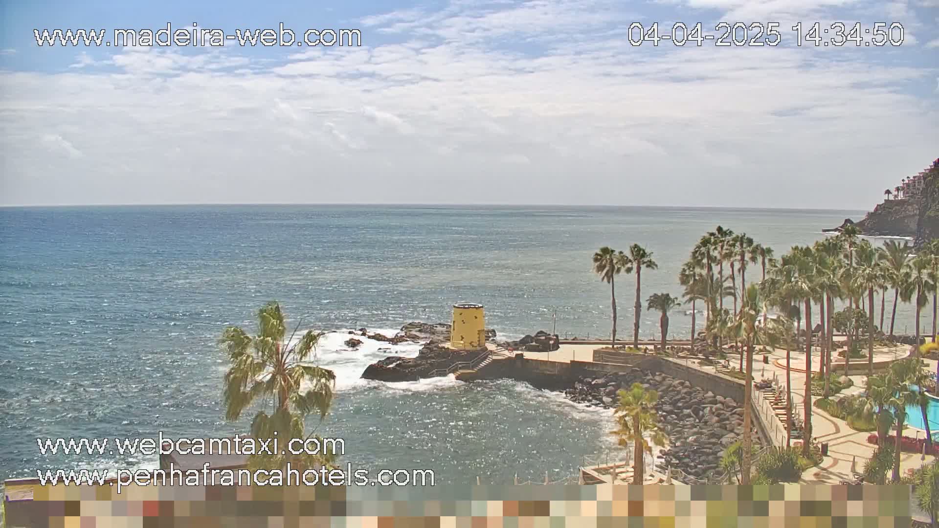 A partly cloudy day reveals a rocky coastline with palm trees and a small yellow tower overlooking a calm ocean.