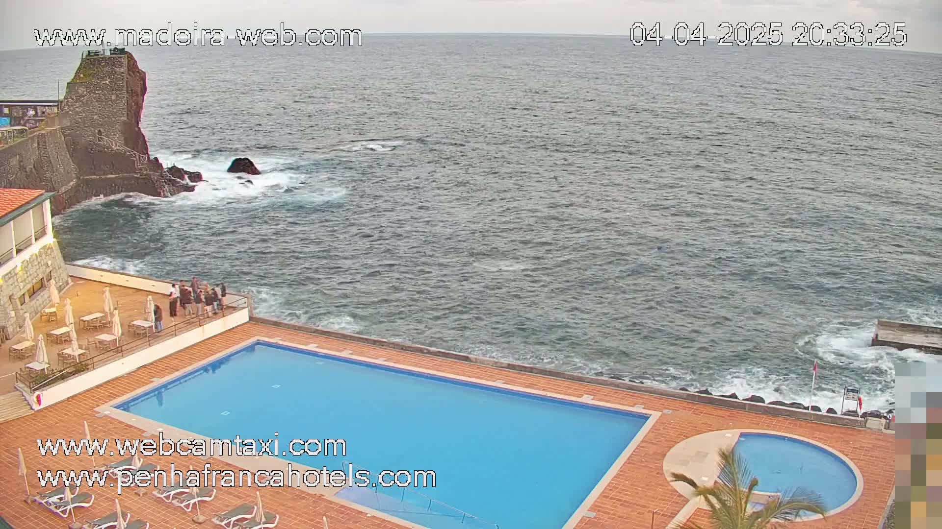 A rectangular swimming pool and a smaller circular pool are situated on a patio overlooking a rocky coastline and a dark, choppy ocean under an overcast sky.