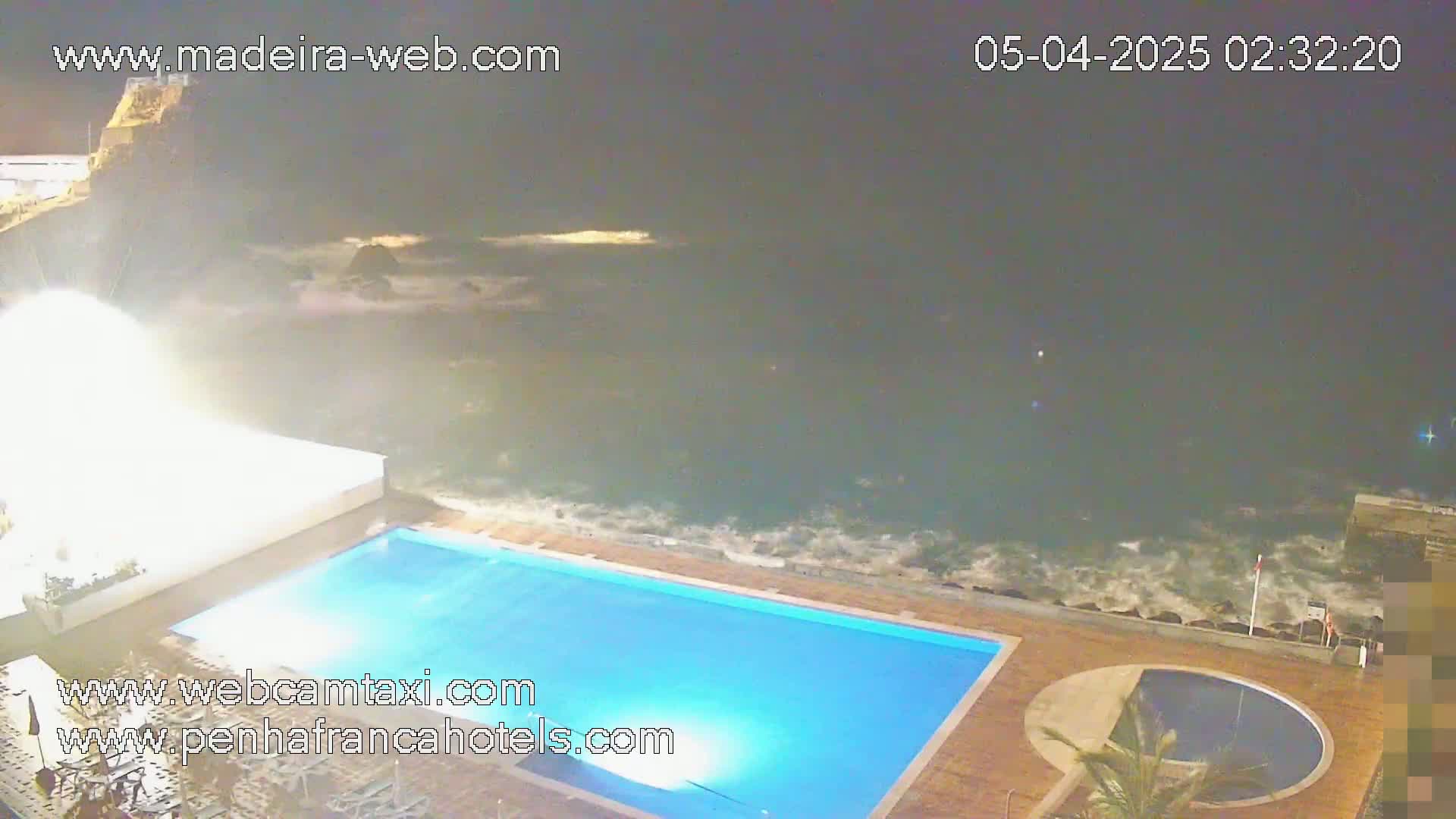 An outdoor swimming pool and a smaller circular pool are next to a rocky ocean shore at night, with waves crashing against the rocks.