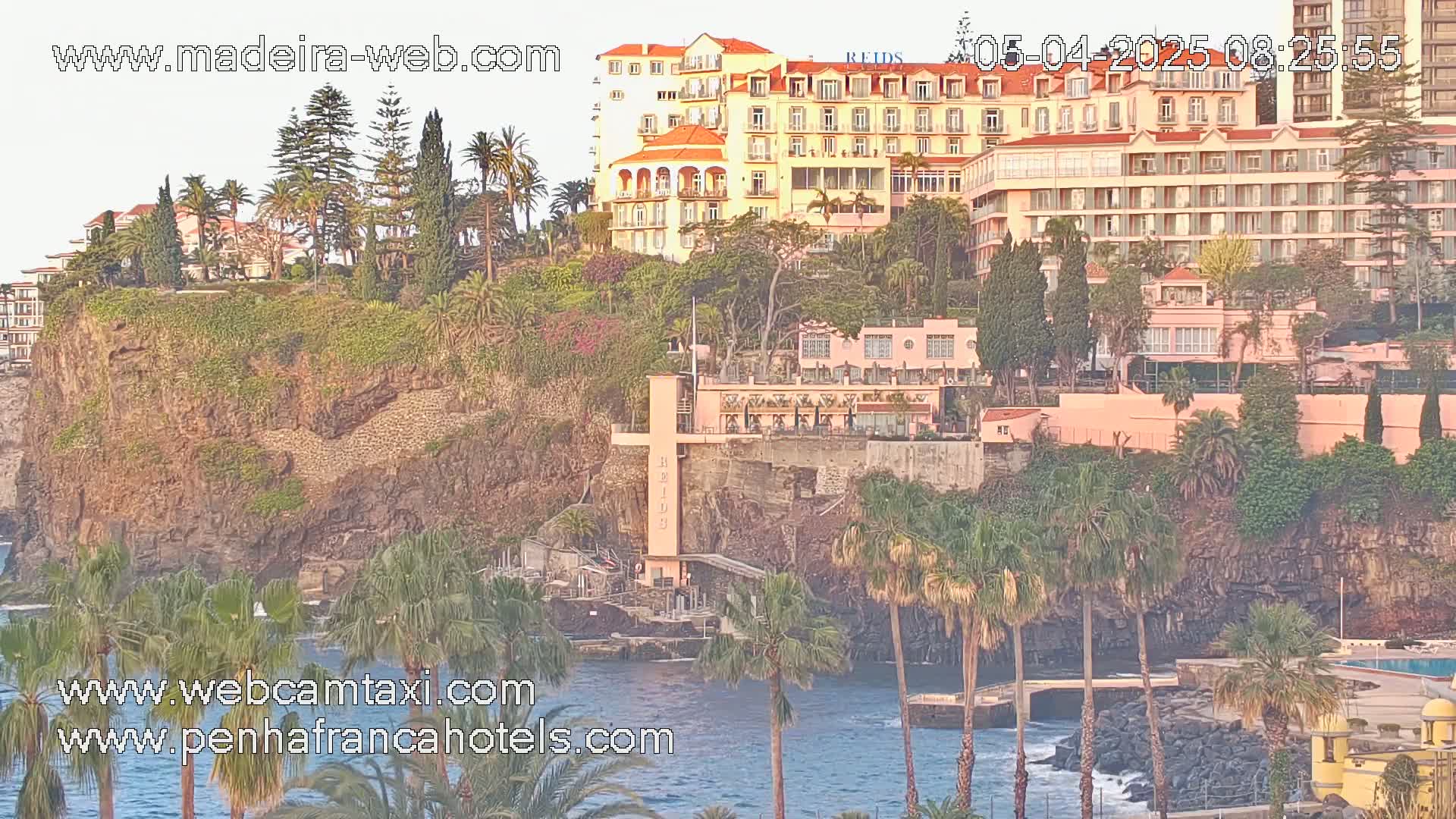A coastal cliffside scene shows several buildings, palm trees, and dark-colored rocky areas with calm water visible below in the soft light of what seems to be either dawn or dusk.