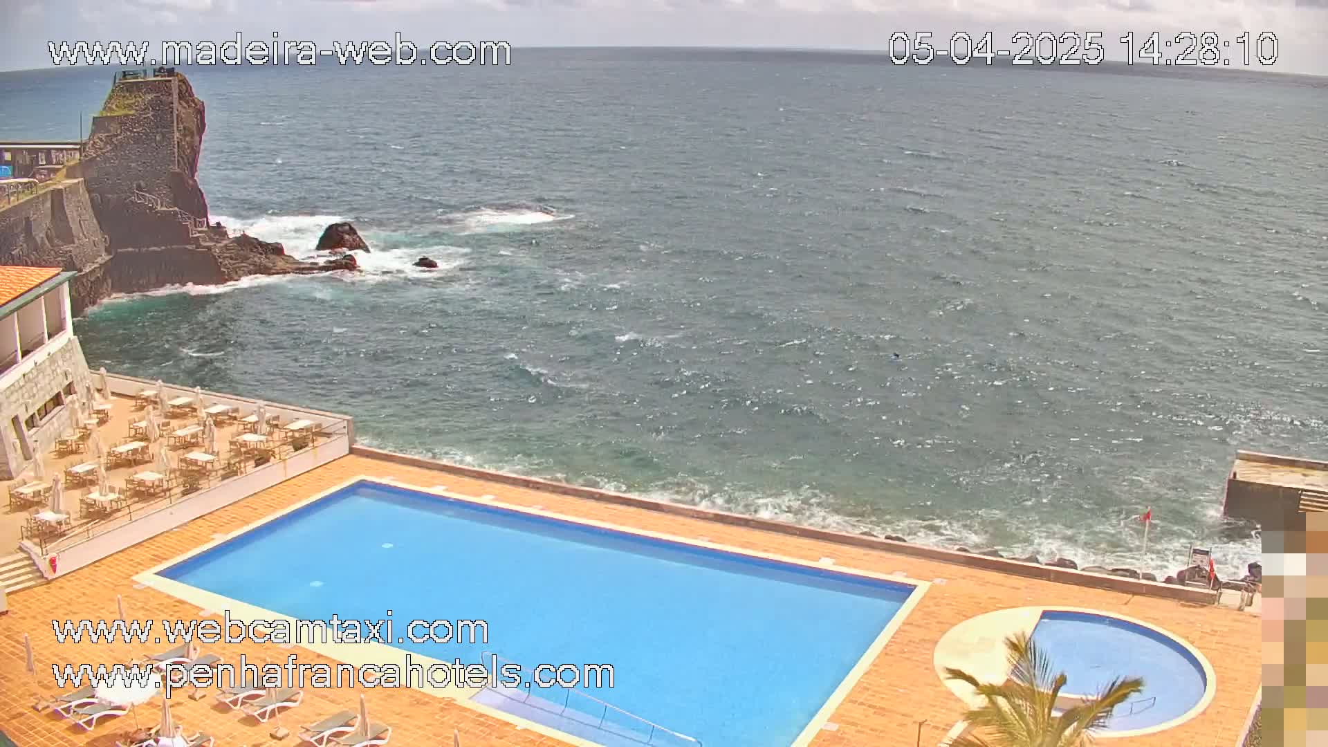 An outdoor swimming pool and a smaller circular pool are situated next to a rocky ocean shore on a partly cloudy day.