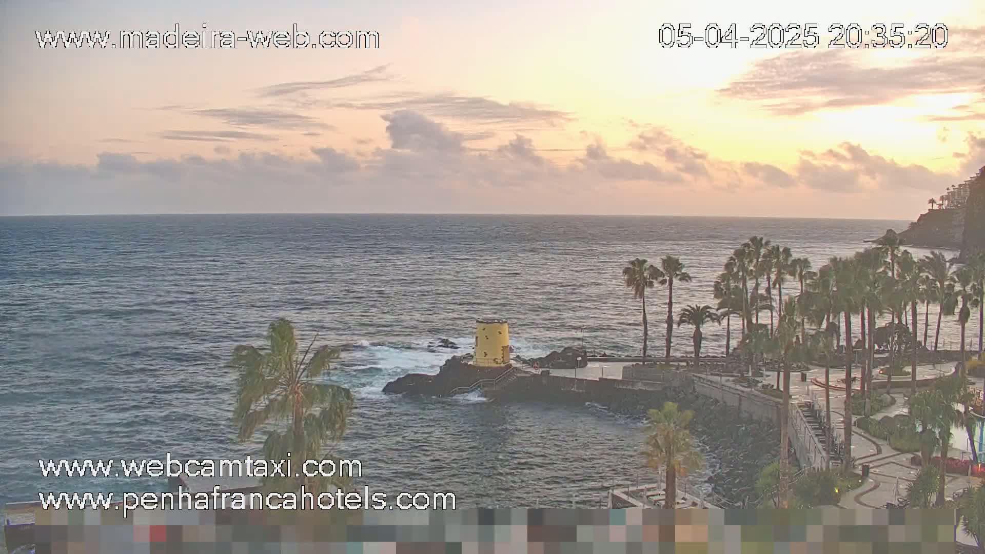 A tranquil ocean scene at sunset, featuring palm trees lining a rocky coastline with a small yellow structure near the water's edge.