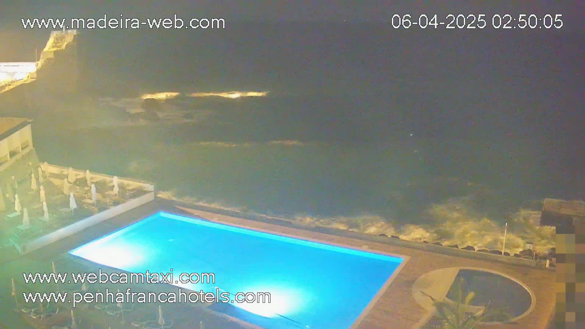 A nighttime view of a swimming pool next to a rocky ocean shore with moderate waves.