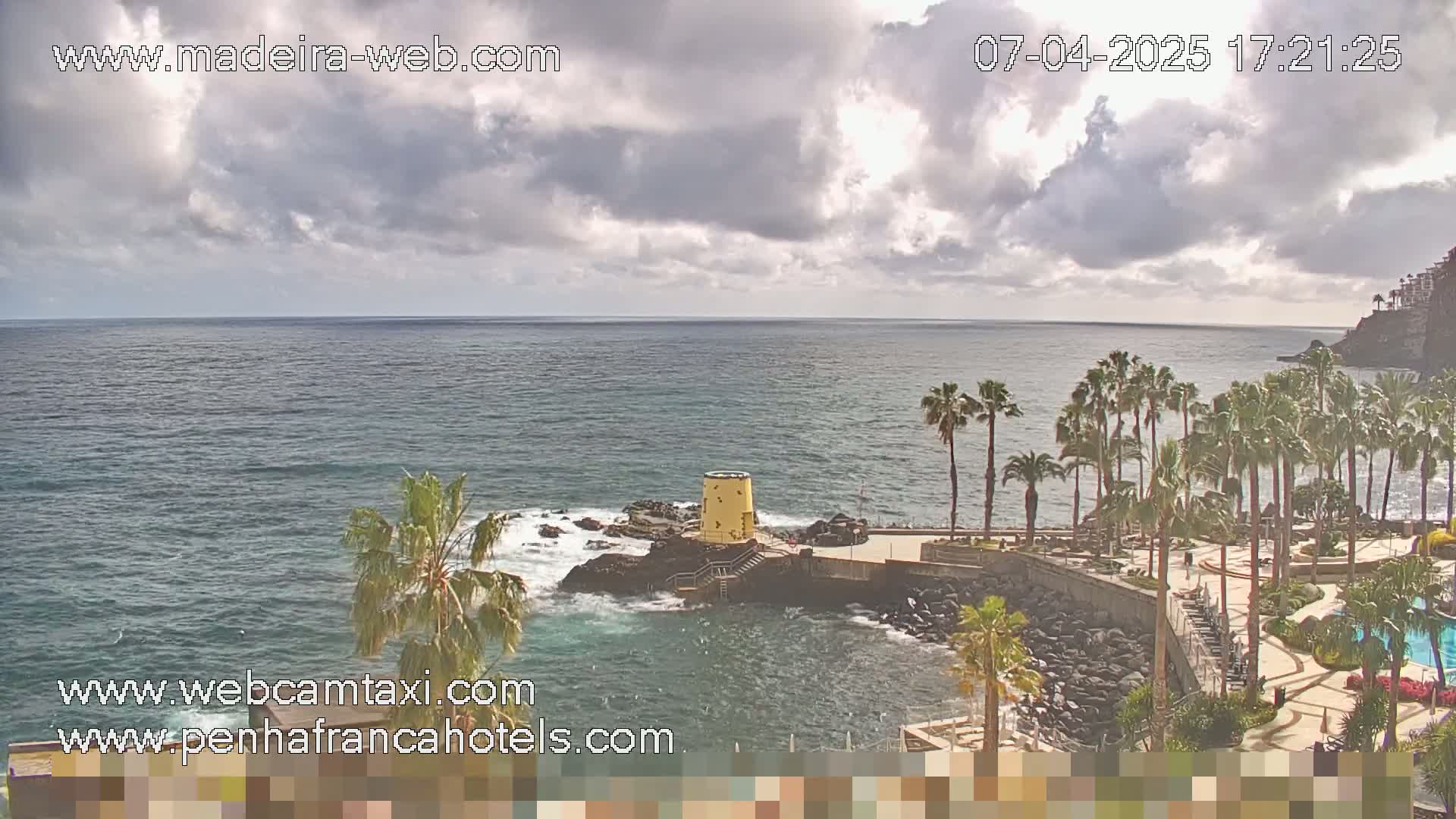 A partly cloudy day shows a rocky coast with palm trees, a small yellow structure near the water, and a calm ocean.