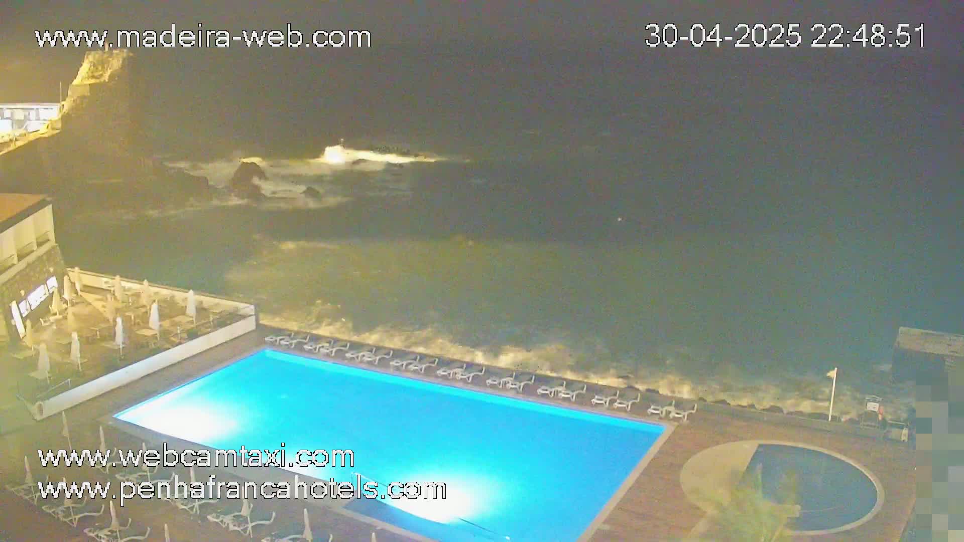 At night, a lighted swimming pool sits adjacent to a rocky ocean shore with crashing waves.