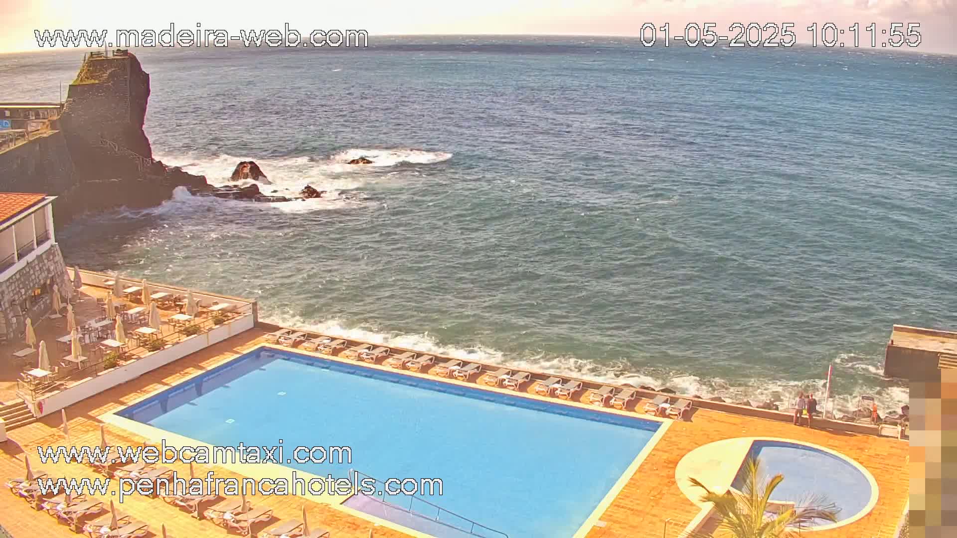 An outdoor swimming pool and lounge area at a seaside resort on a partly sunny day with somewhat choppy water.