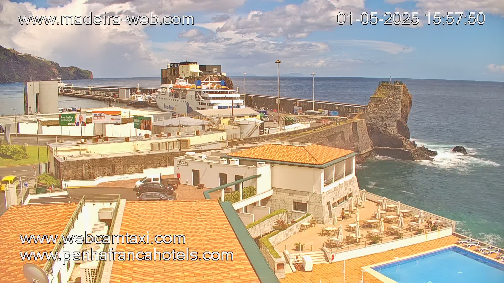 A partly cloudy day shows a harbor with a ferry docked, buildings along a rocky coast, and a hotel with an outdoor pool and patio seating area.