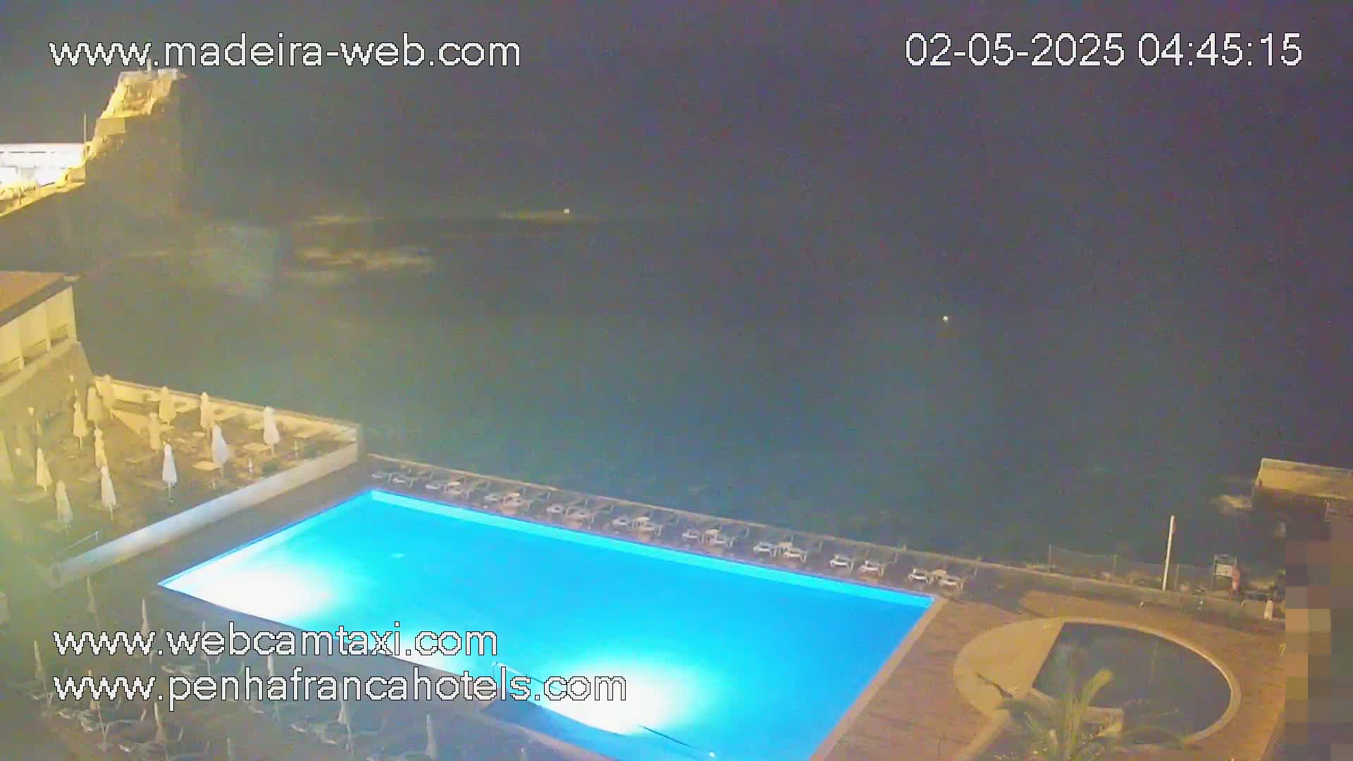A nighttime view of a brightly lit, rectangular swimming pool on a patio overlooking a dark ocean.