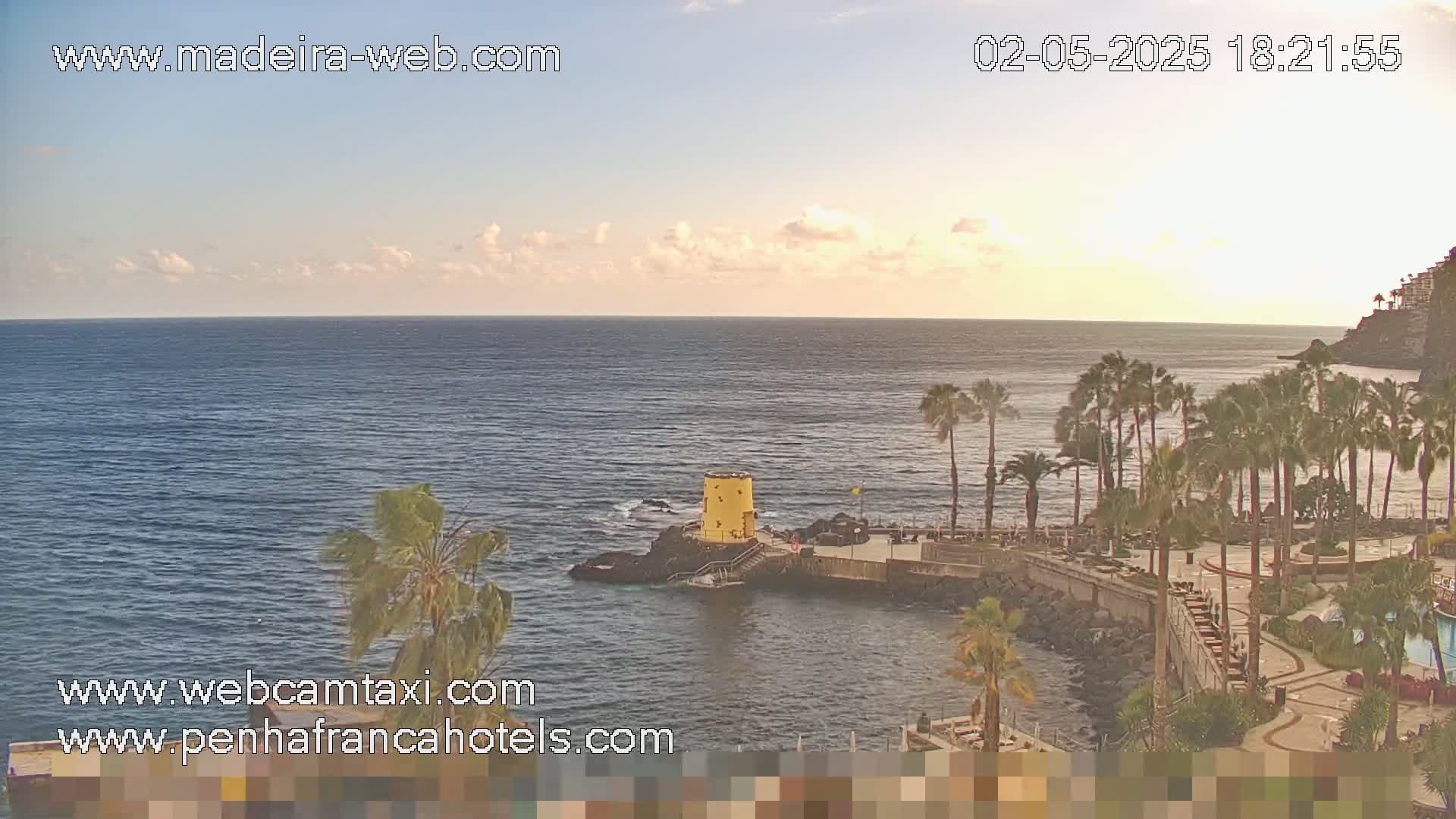 A calm ocean under a partly cloudy sunset sky is viewed from a resort with palm trees and a small yellow tower on a rocky point.