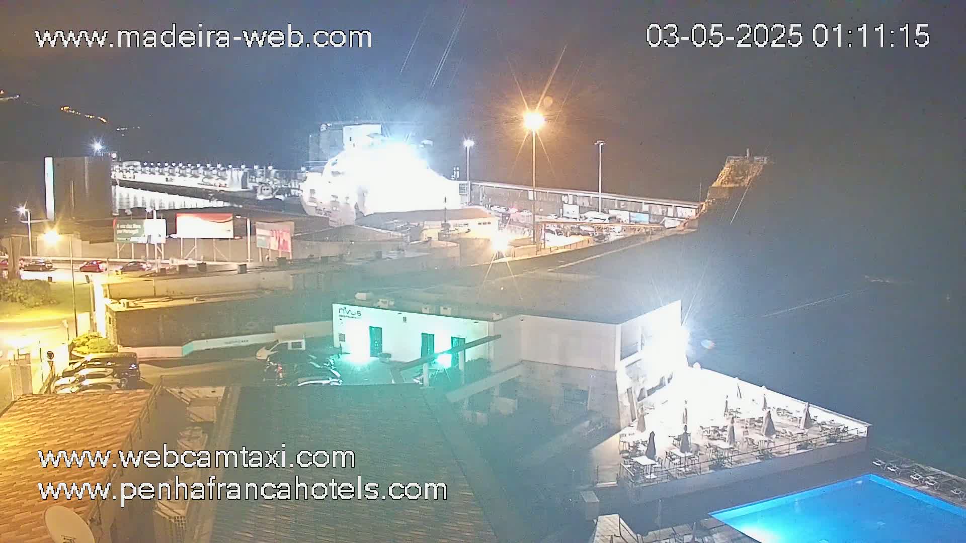 A nighttime aerial view shows a brightly lit waterfront area with a large building, a pool, and parked cars, under clear skies.