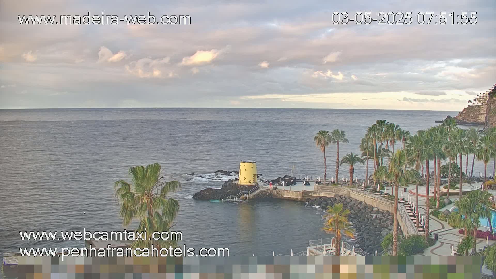 A partly cloudy sky overlooks a calm ocean with palm trees lining a rocky coast next to a resort area.
