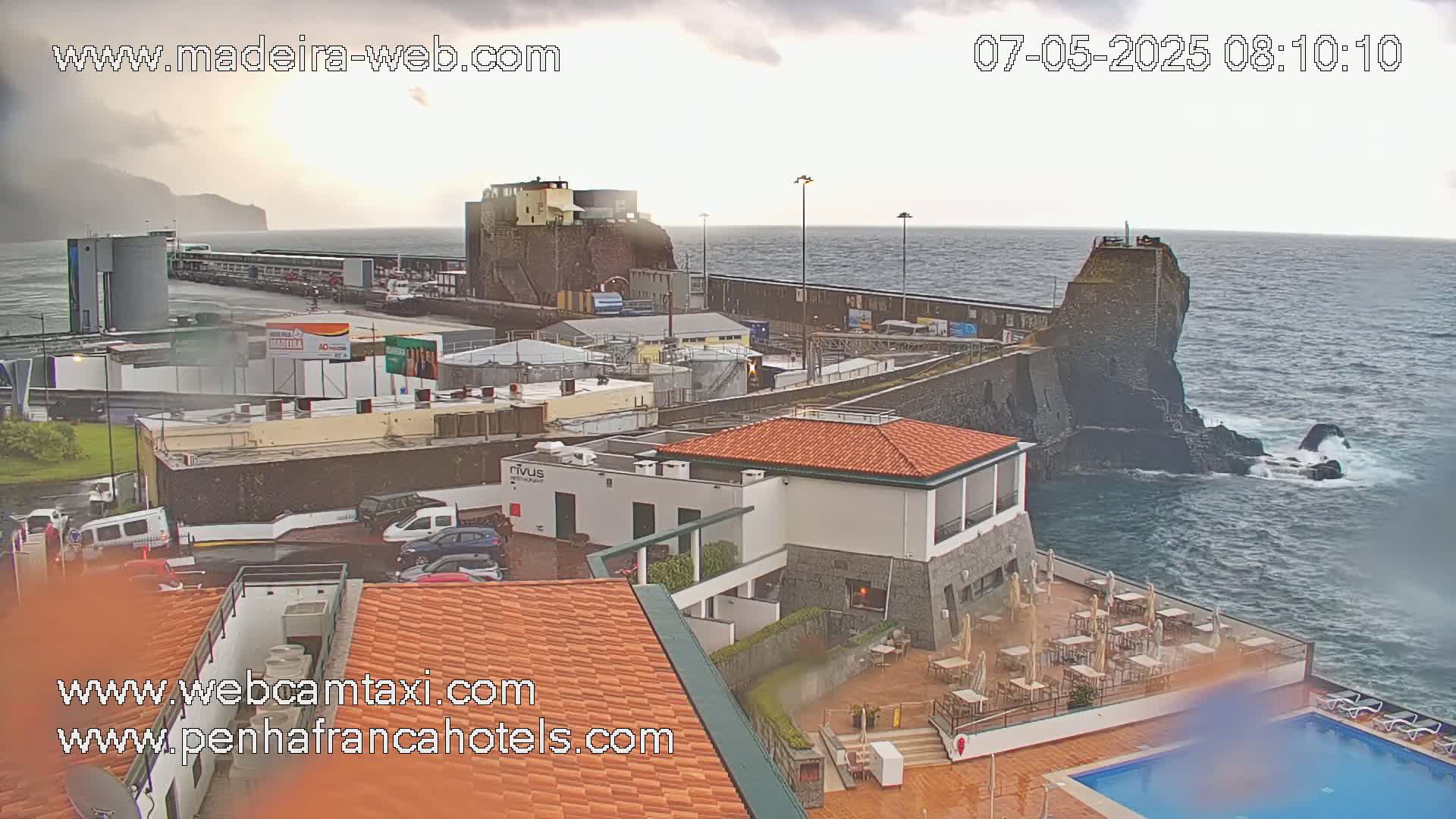 An overcast day shows a coastal scene with a rocky outcrop, buildings, a parking lot, and an outdoor pool area, all overlooking a choppy ocean.