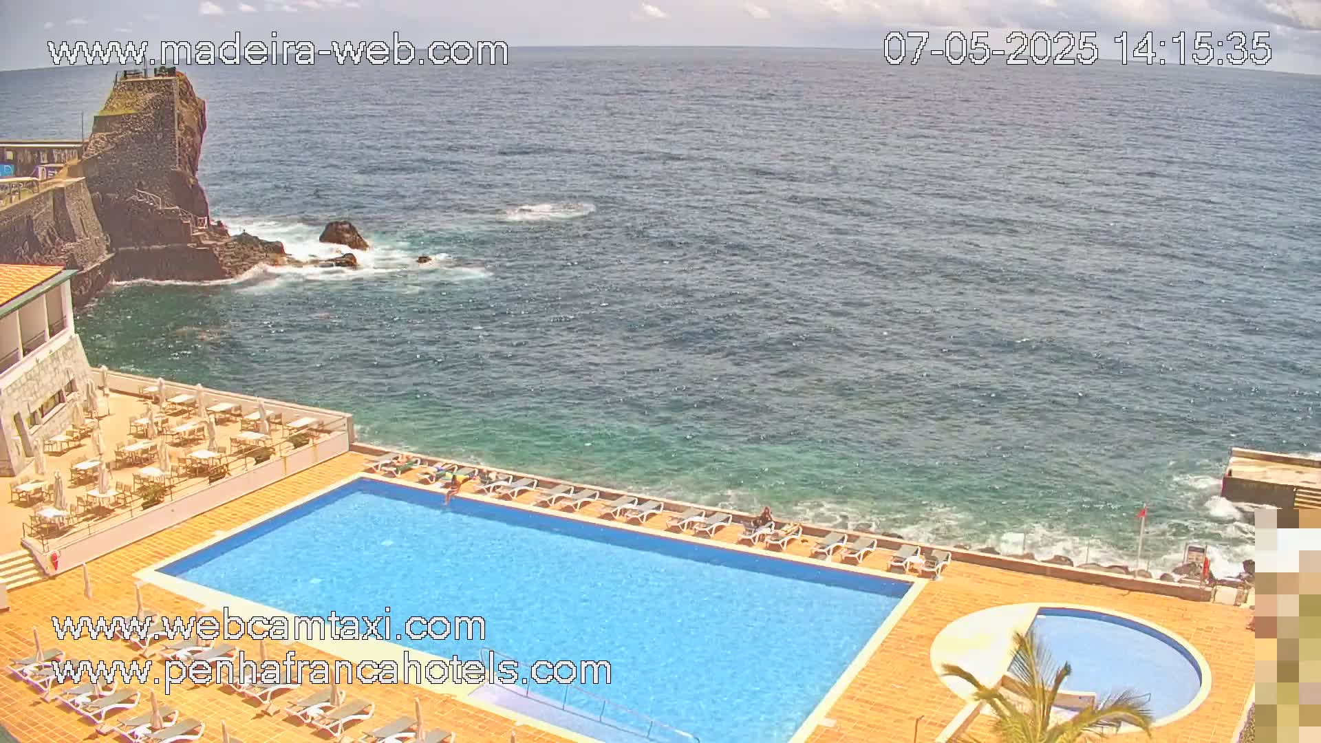 An outdoor swimming pool and smaller circular pool are situated on a cliff overlooking a calm ocean on a partly cloudy day.