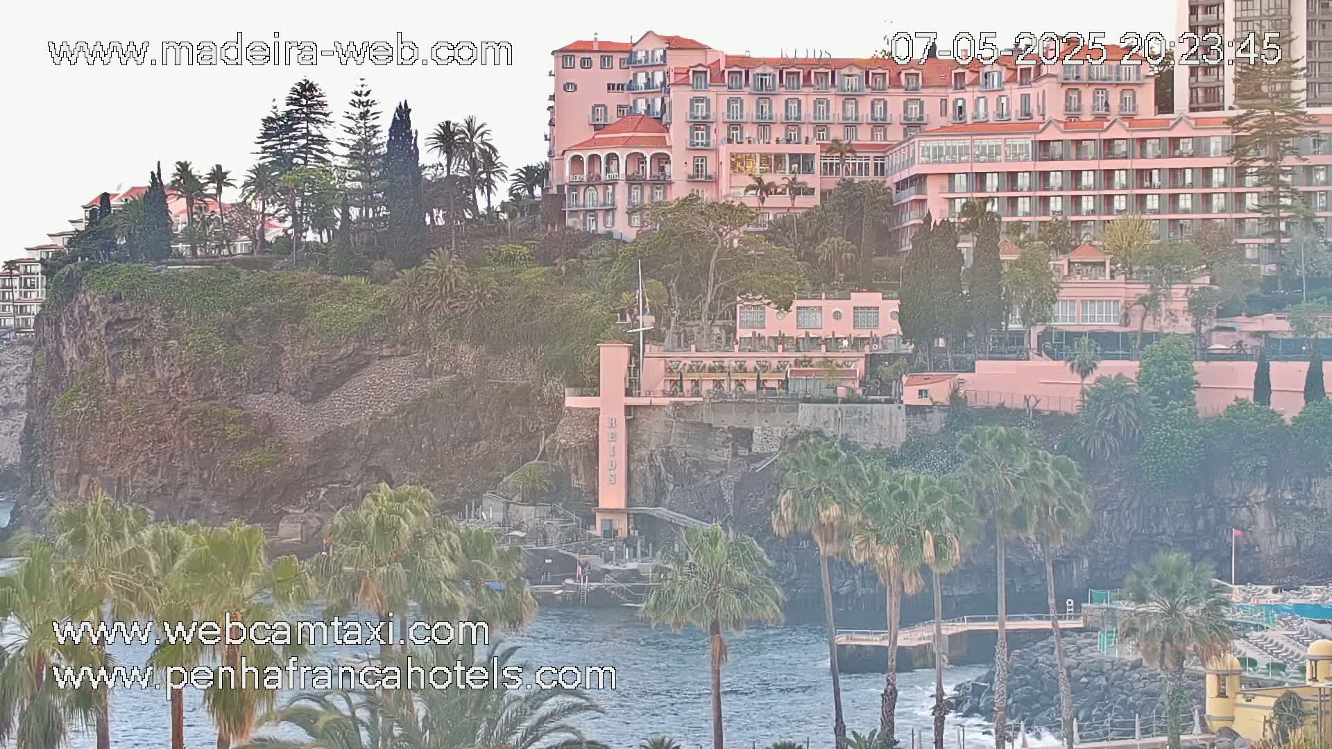 A pink hotel is situated on a cliff overlooking a calm ocean, surrounded by lush greenery and palm trees.