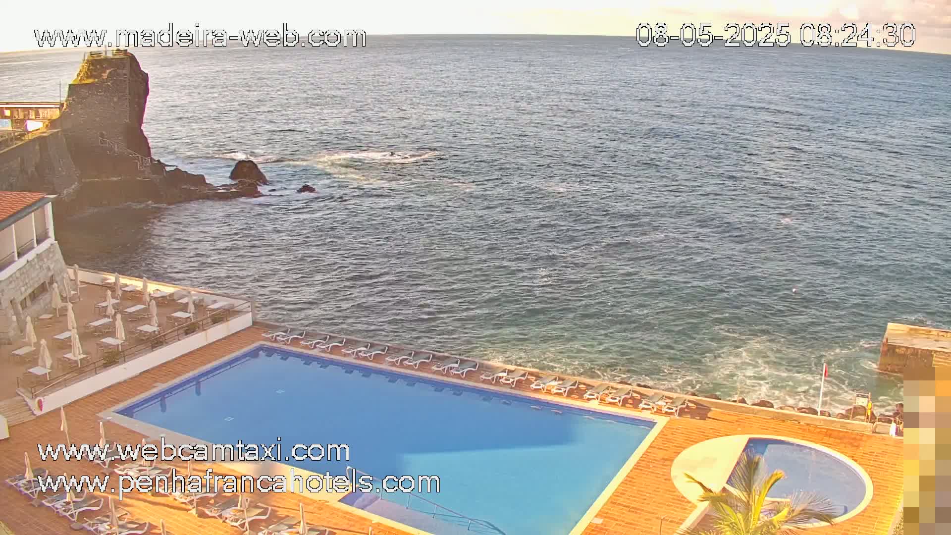 An outdoor swimming pool and sun deck area overlooks a calm ocean with a rocky shoreline and a small stone structure visible in the distance.