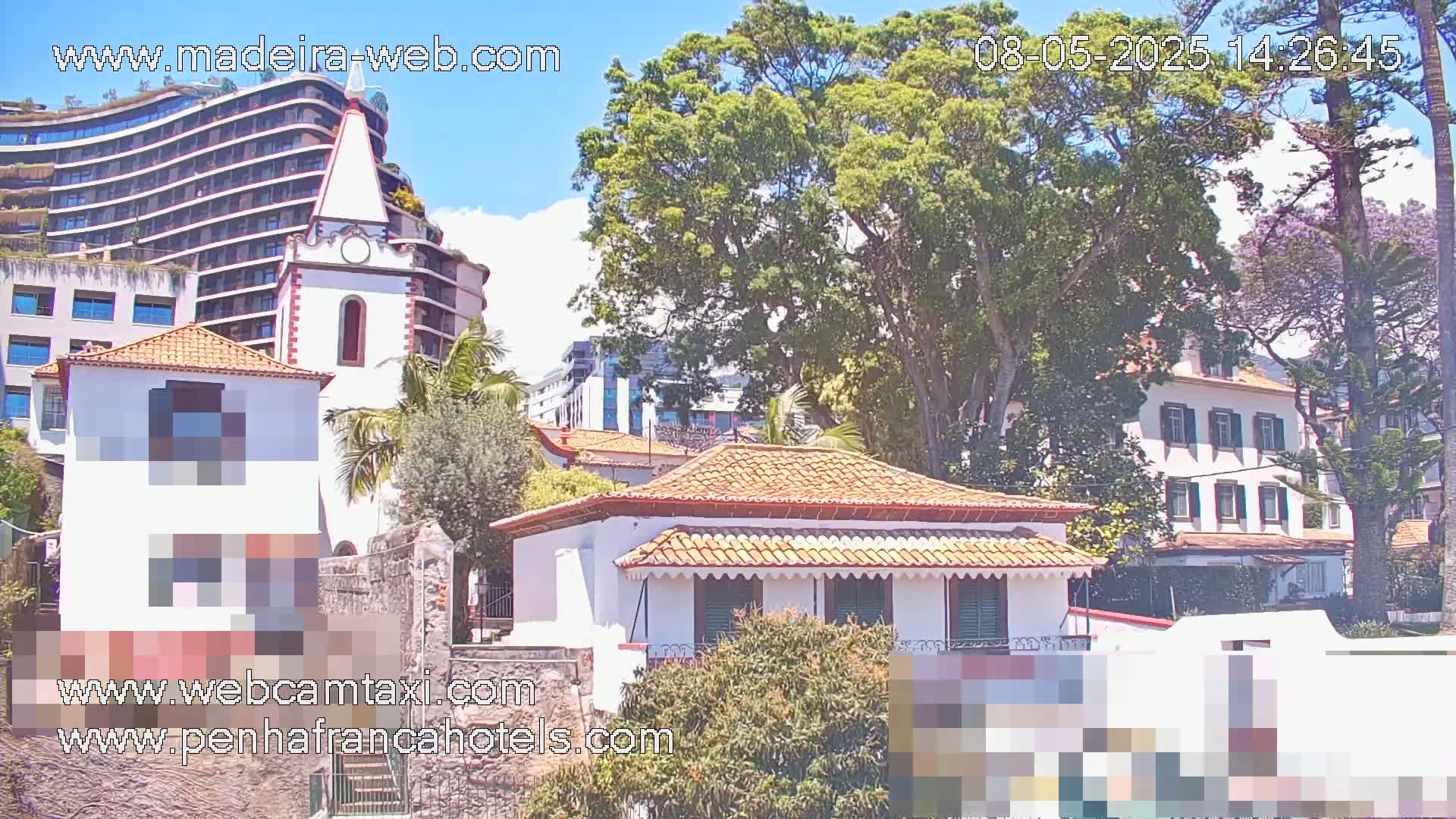 A sunny day reveals a cluster of buildings, including a whitewashed house with a terracotta roof, a church tower, and a modern high-rise structure, nestled amongst lush green trees.