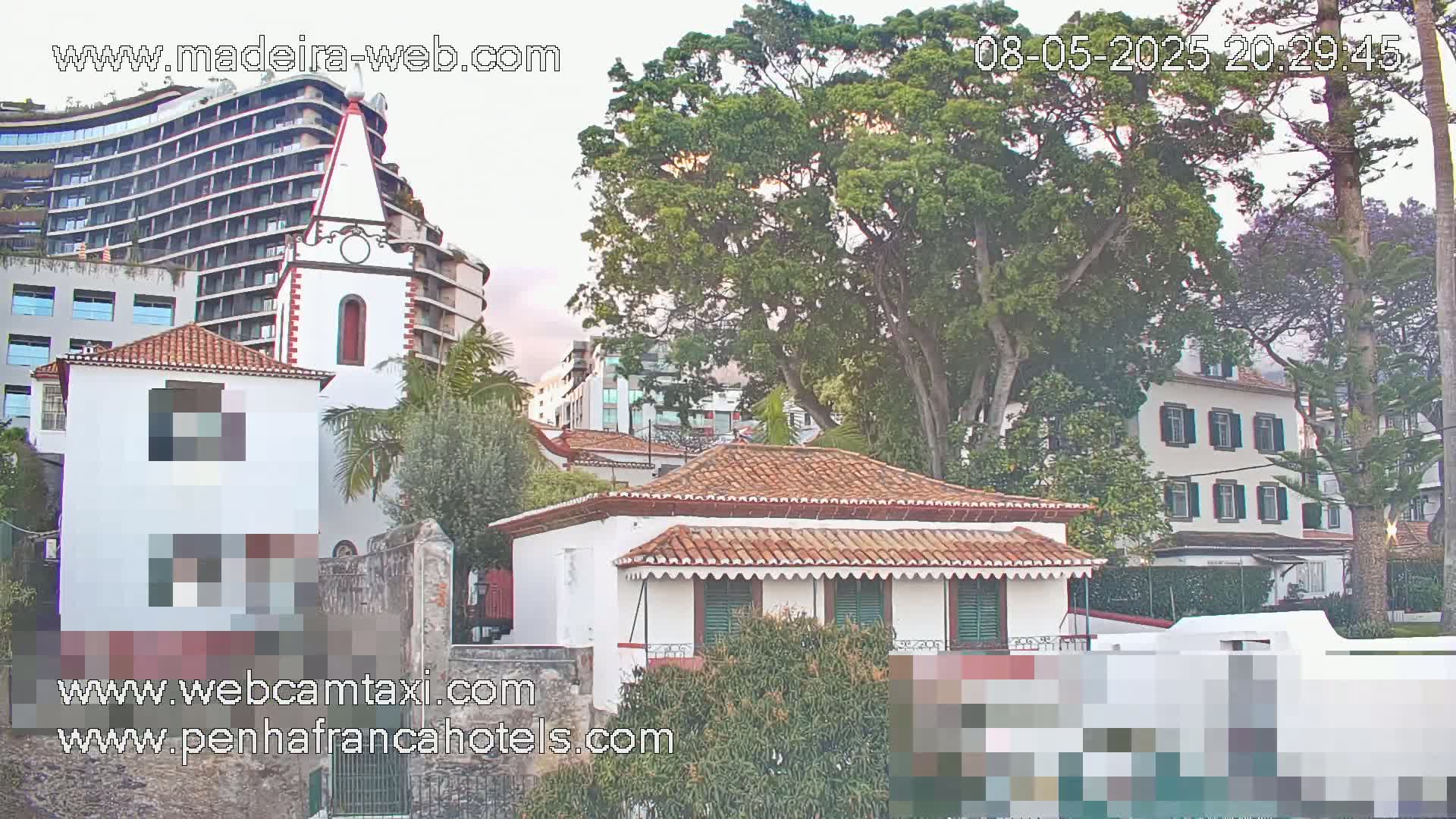 A white building with a red tile roof stands next to a taller white church, all surrounded by lush green trees under an overcast sky.