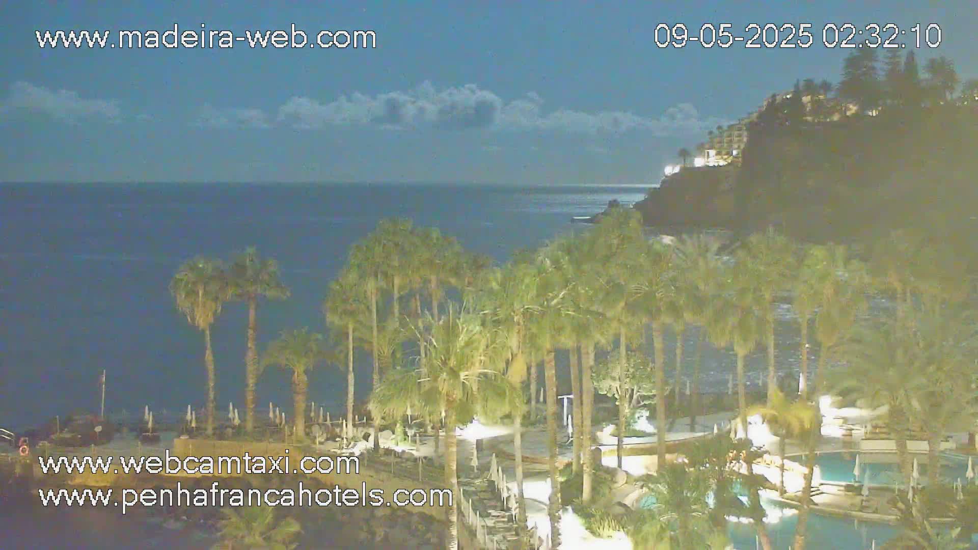 A nighttime view of a resort with palm trees illuminated near a dark ocean under a partly cloudy sky.