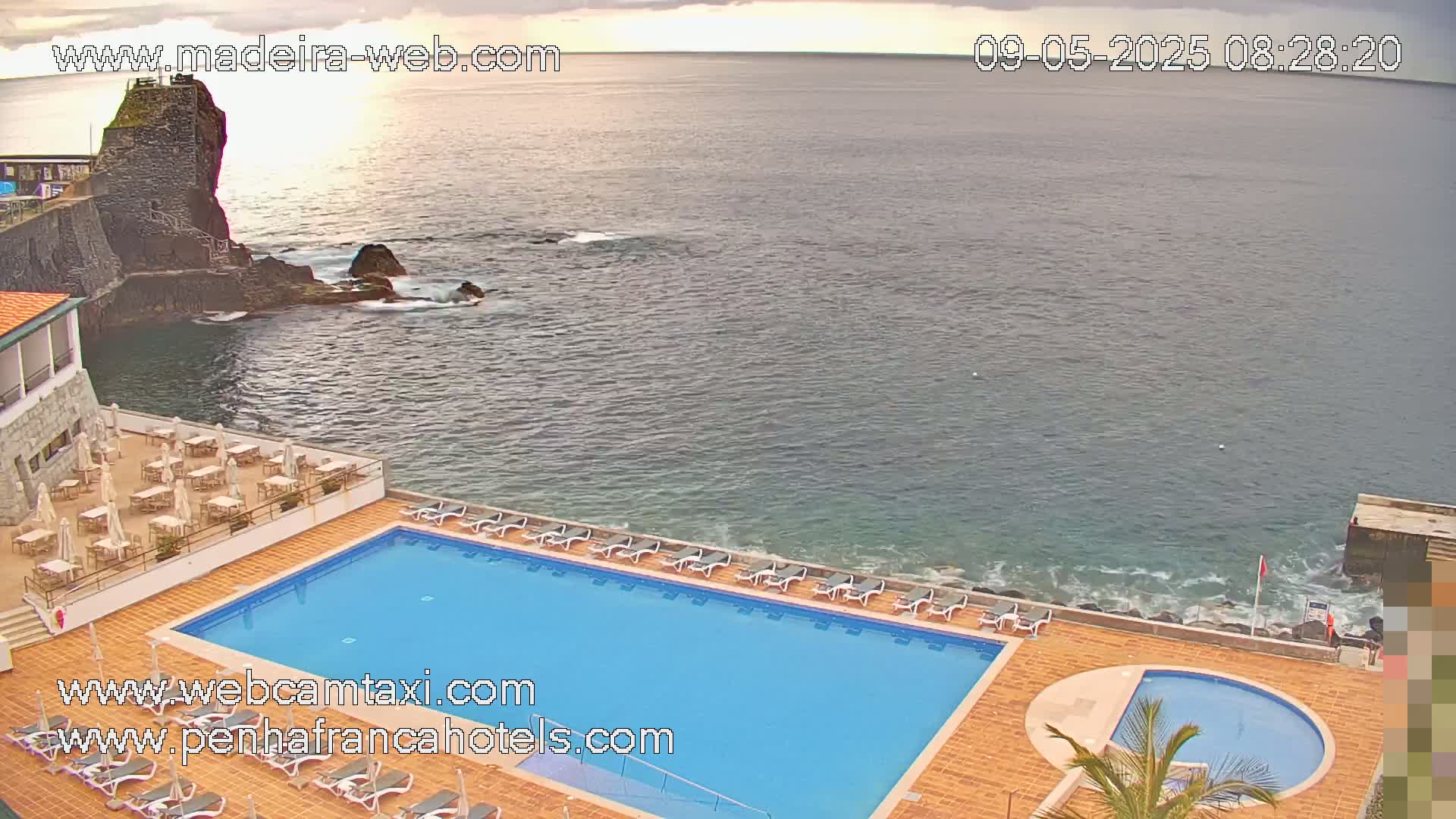 An outdoor swimming pool and lounge area at a hotel overlooks a calm ocean next to a rocky outcropping under a partly cloudy sky.