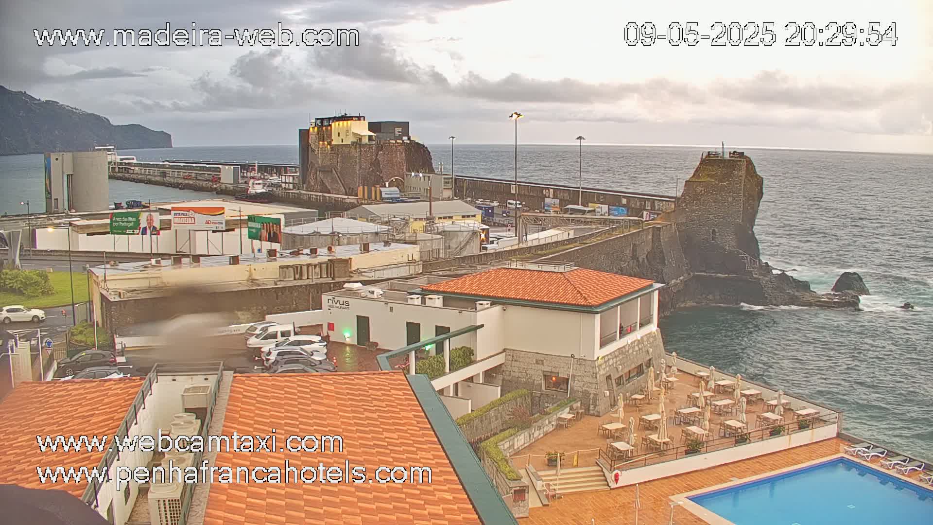 An overcast day shows a coastal town with a harbor, a stone fortress, a hotel with an outdoor pool and patio, and the ocean with waves crashing against the rocks.