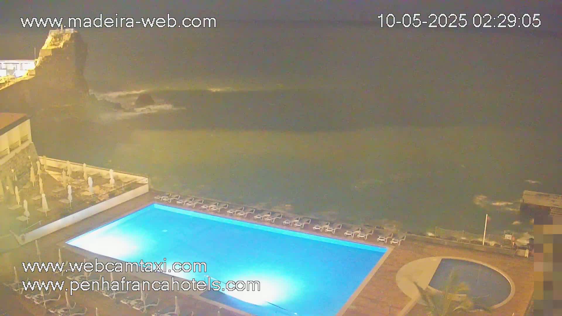 An illuminated swimming pool at night overlooks a dark ocean with small waves under a dark sky.