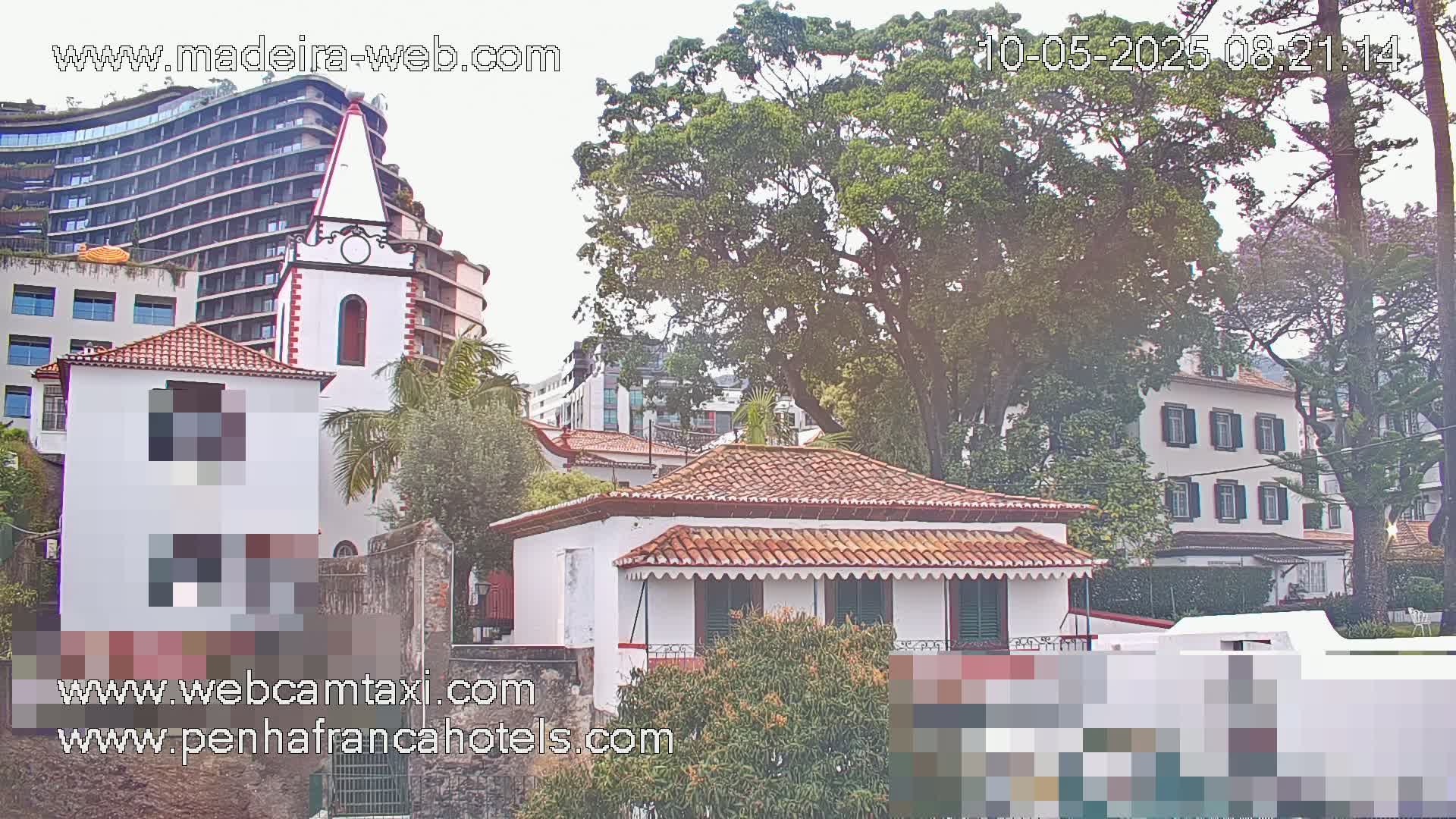 A view of buildings, including a church and a small house with red tile roofs, partially obscured by trees, under an overcast sky.