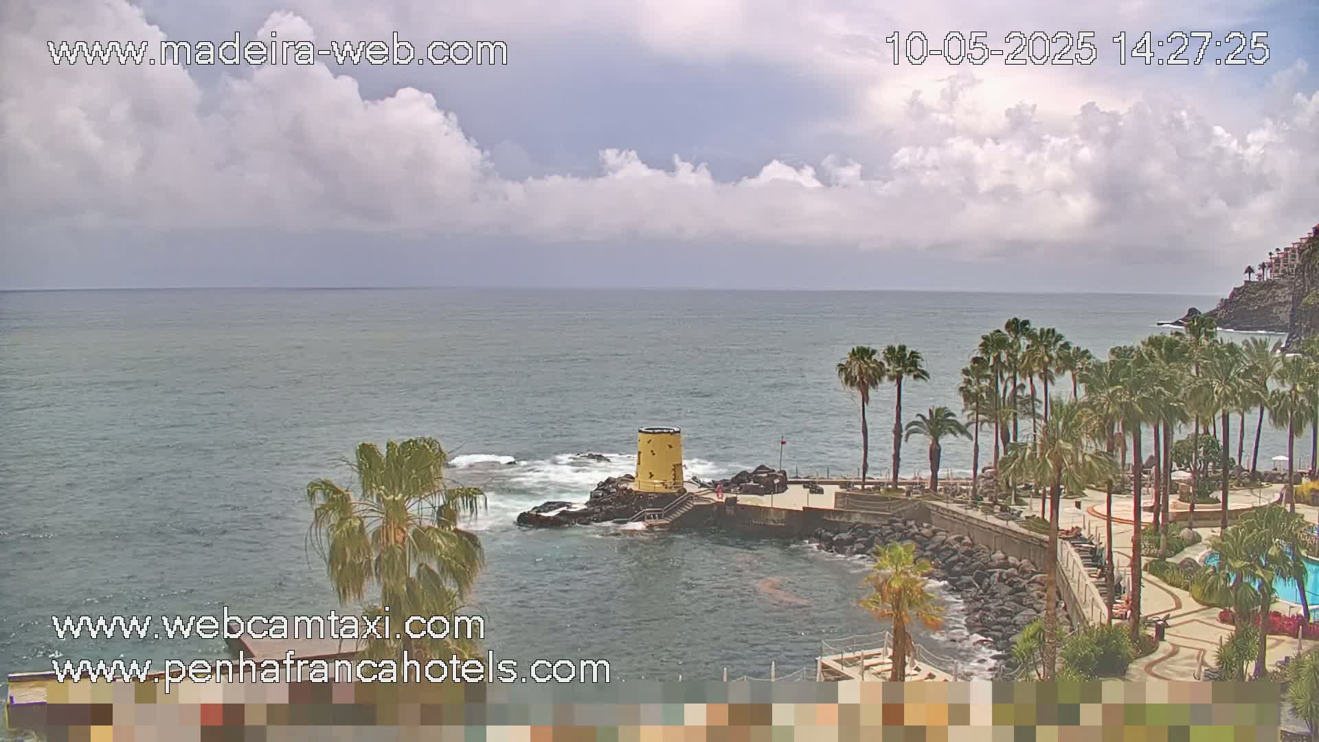 A partly cloudy day shows a rocky coast with palm trees and a small yellow tower overlooking a calm ocean.