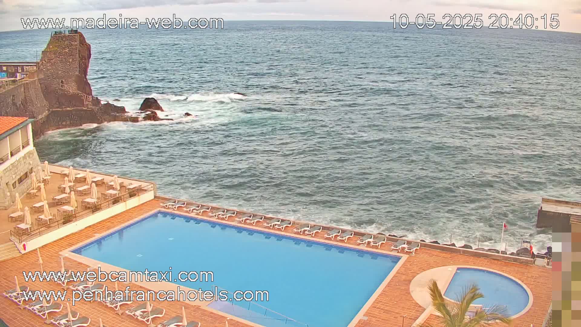 An outdoor swimming pool and lounge area are situated next to a rocky coastline with a partly cloudy sky and moderate waves.