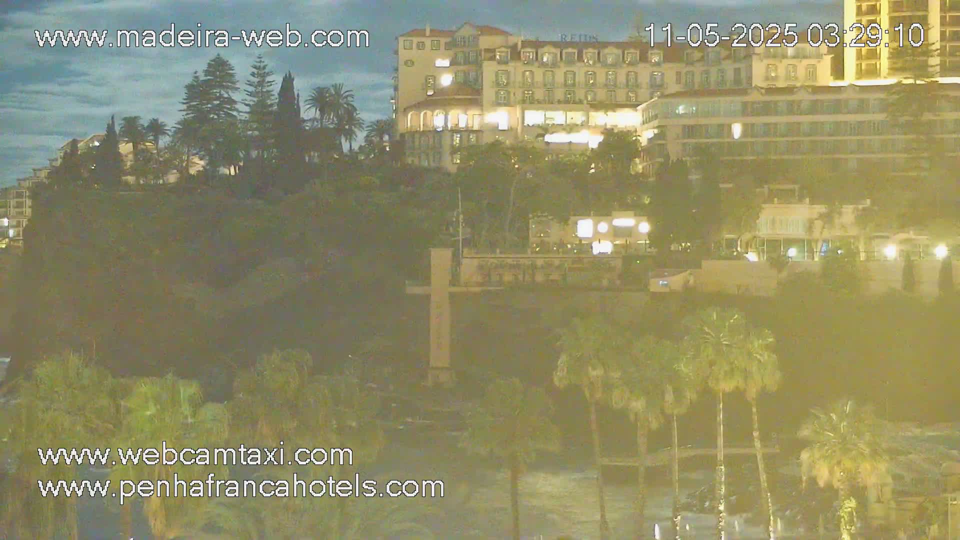 A nighttime view of illuminated buildings and palm trees on a hillside overlooking a dark body of water under a cloudy sky.
