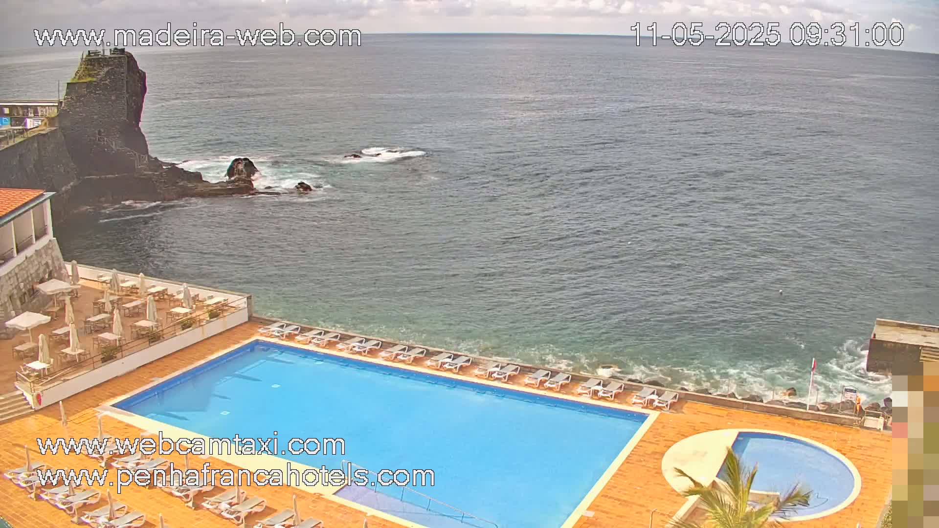 An outdoor pool with lounge chairs sits beside a rocky ocean shore on an overcast day.