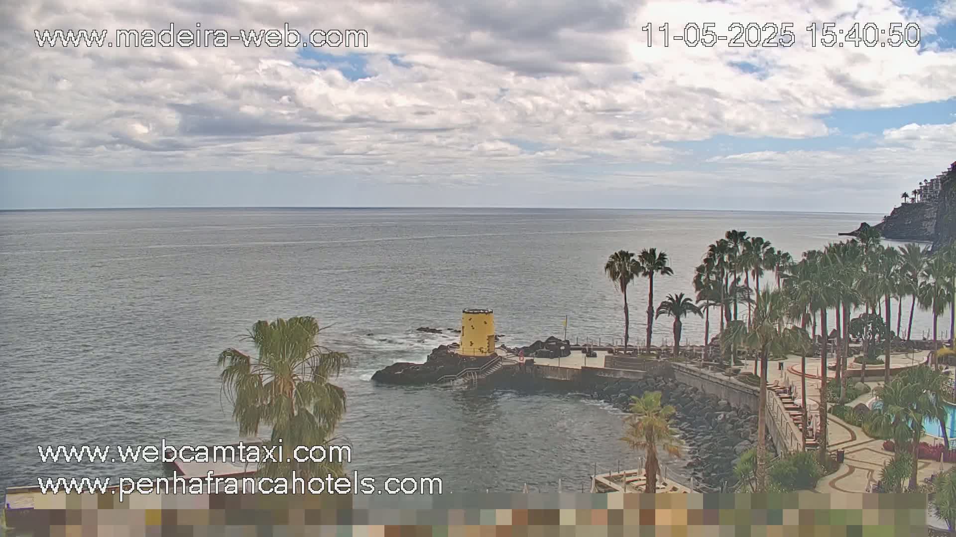 A partly cloudy day shows a rocky coastline with palm trees, a yellow tower, and calm ocean water.