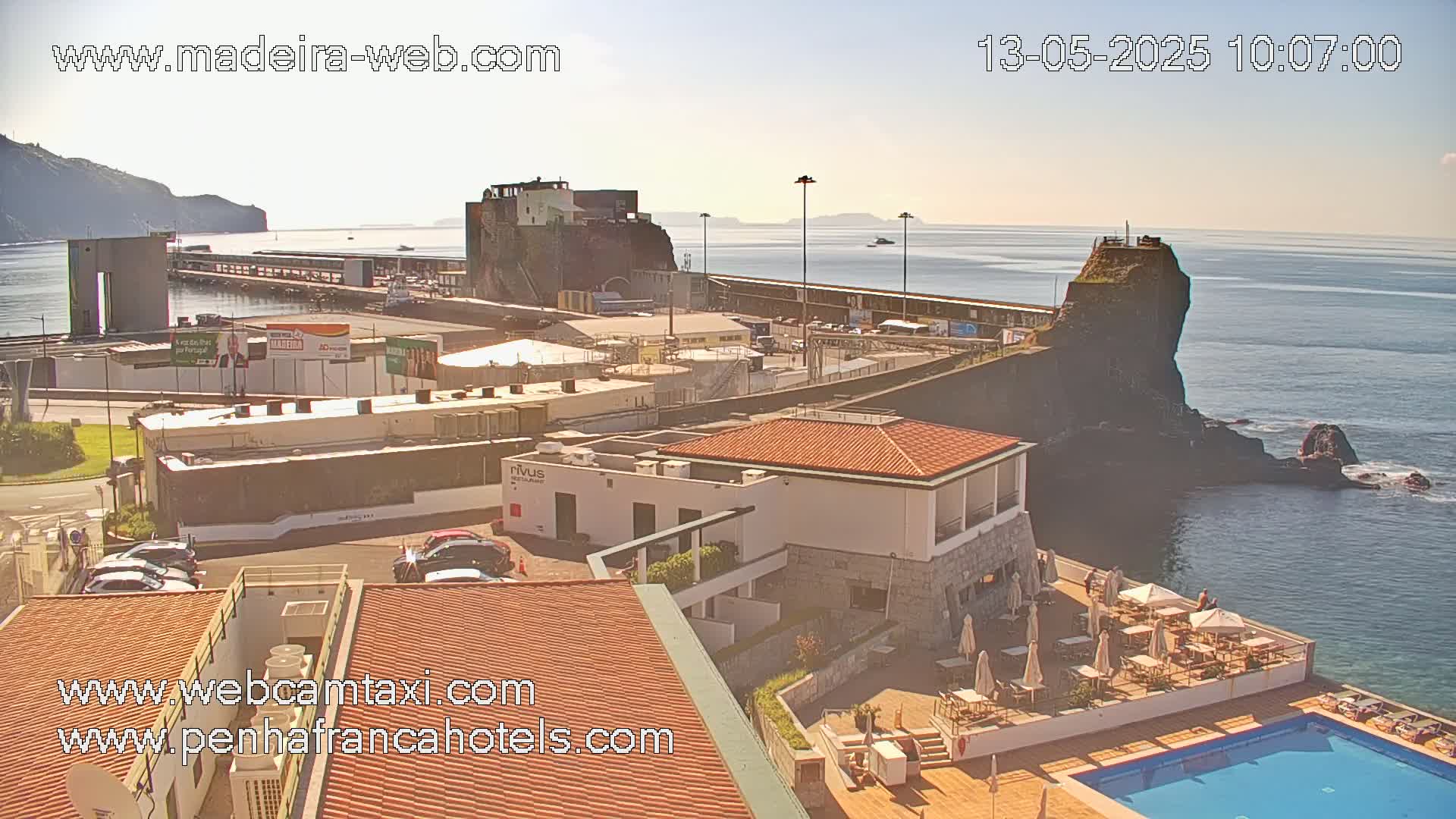 A sunny coastal scene shows a harbor with buildings, a rocky outcrop, and a hotel with an outdoor pool.