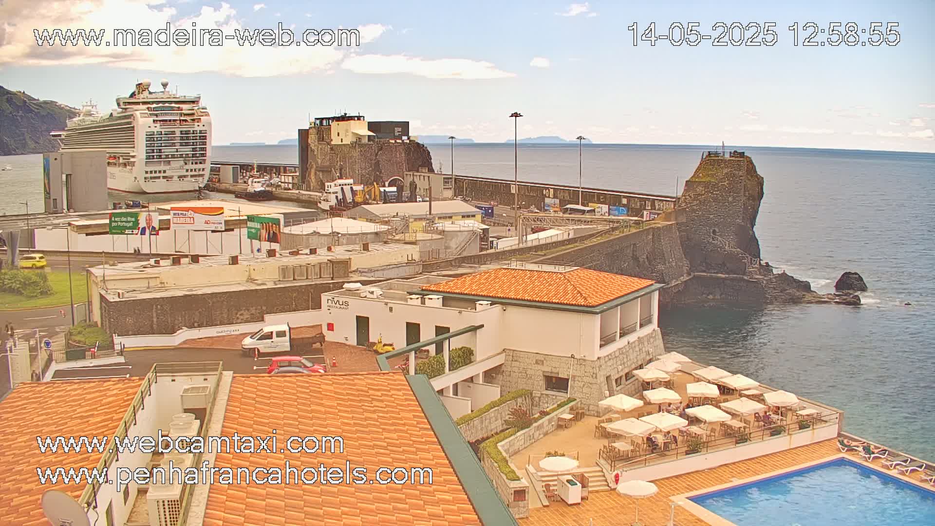 A cruise ship is docked at a harbor next to a stone building and a hotel with an outdoor pool and patio under a partly cloudy sky.