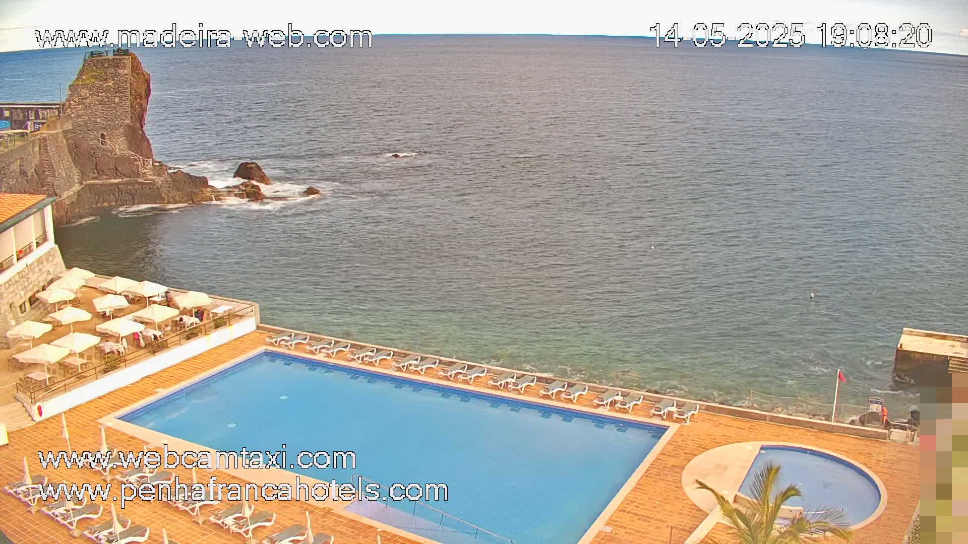 A resort pool and patio area with lounge chairs overlooks a calm ocean next to a rocky shoreline with a stone structure.