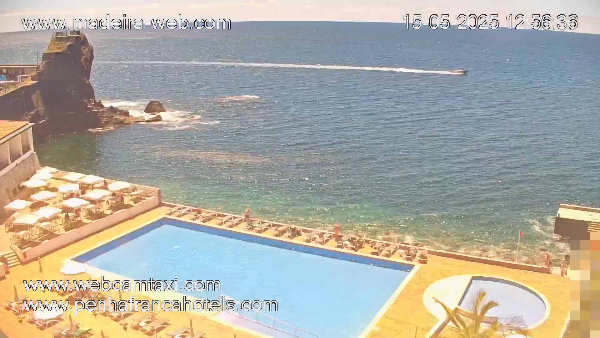 A sunny day overlooks a resort's pool area next to a rocky ocean shoreline with a boat in the distance.