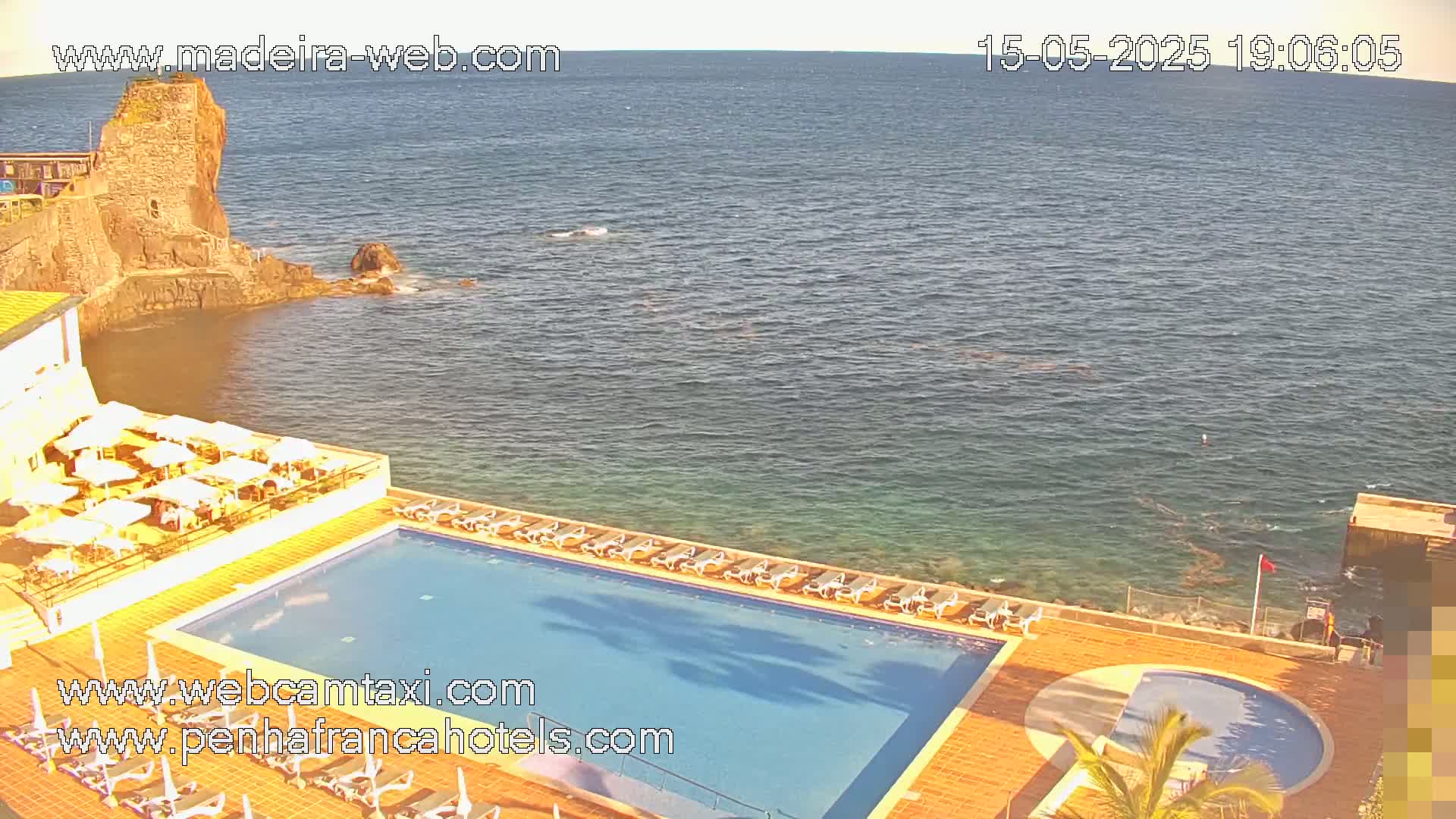 An outdoor pool and patio area at a hotel overlook a calm ocean next to a rocky shoreline with a stone structure.