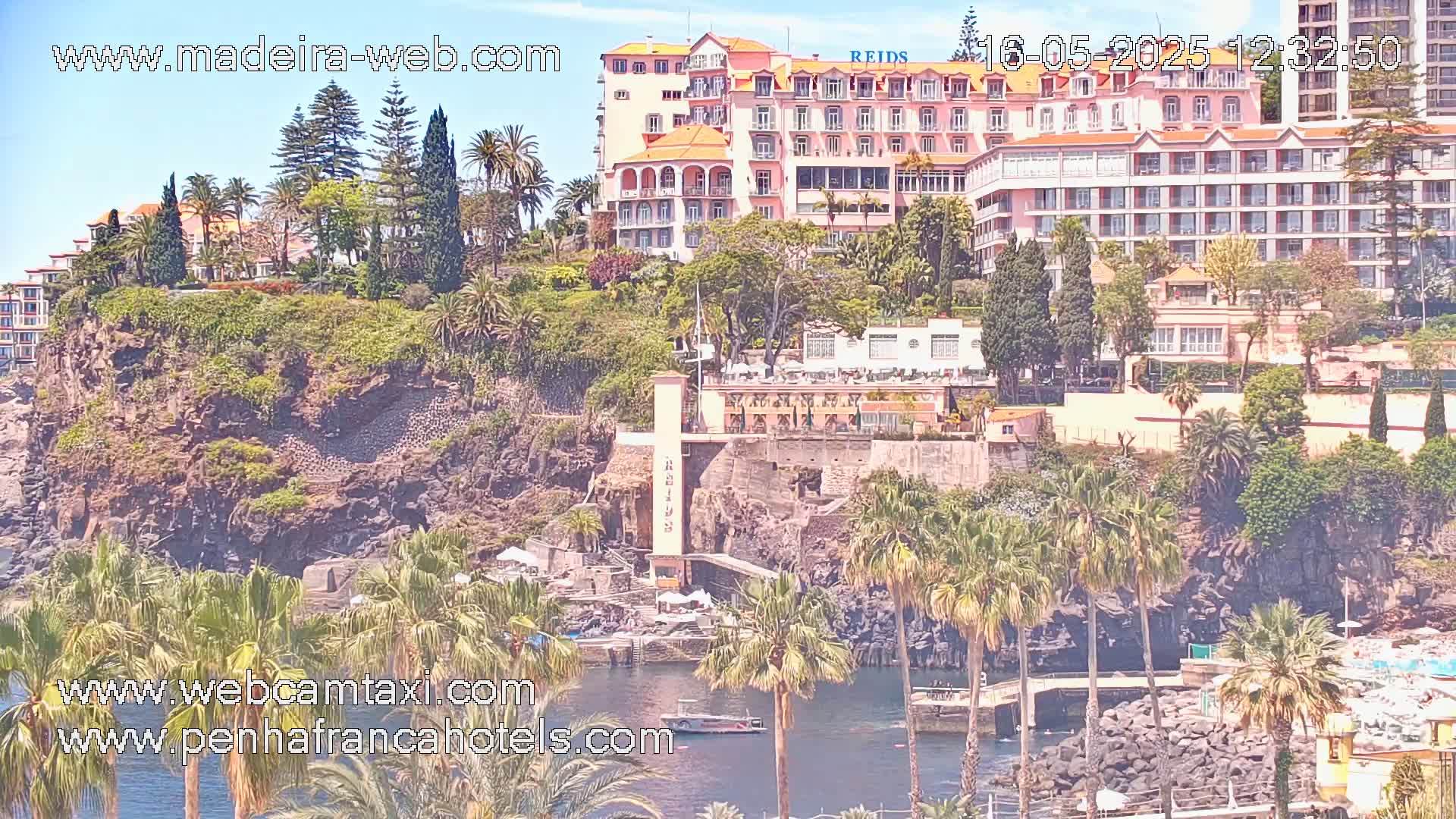 A sunny coastal scene features a large pink hotel perched atop a rocky cliff, surrounded by lush tropical vegetation and palm trees, overlooking a calm body of water.