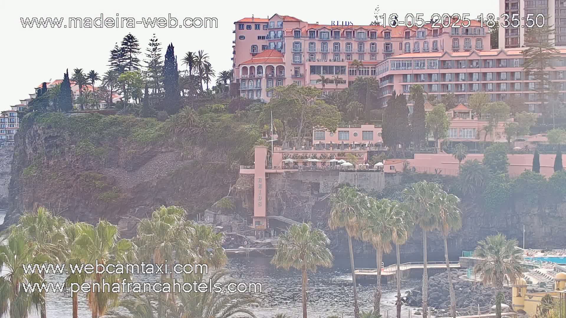 A hazy view of a large pink hotel complex situated on a cliff overlooking a calm body of water, surrounded by lush vegetation and palm trees.