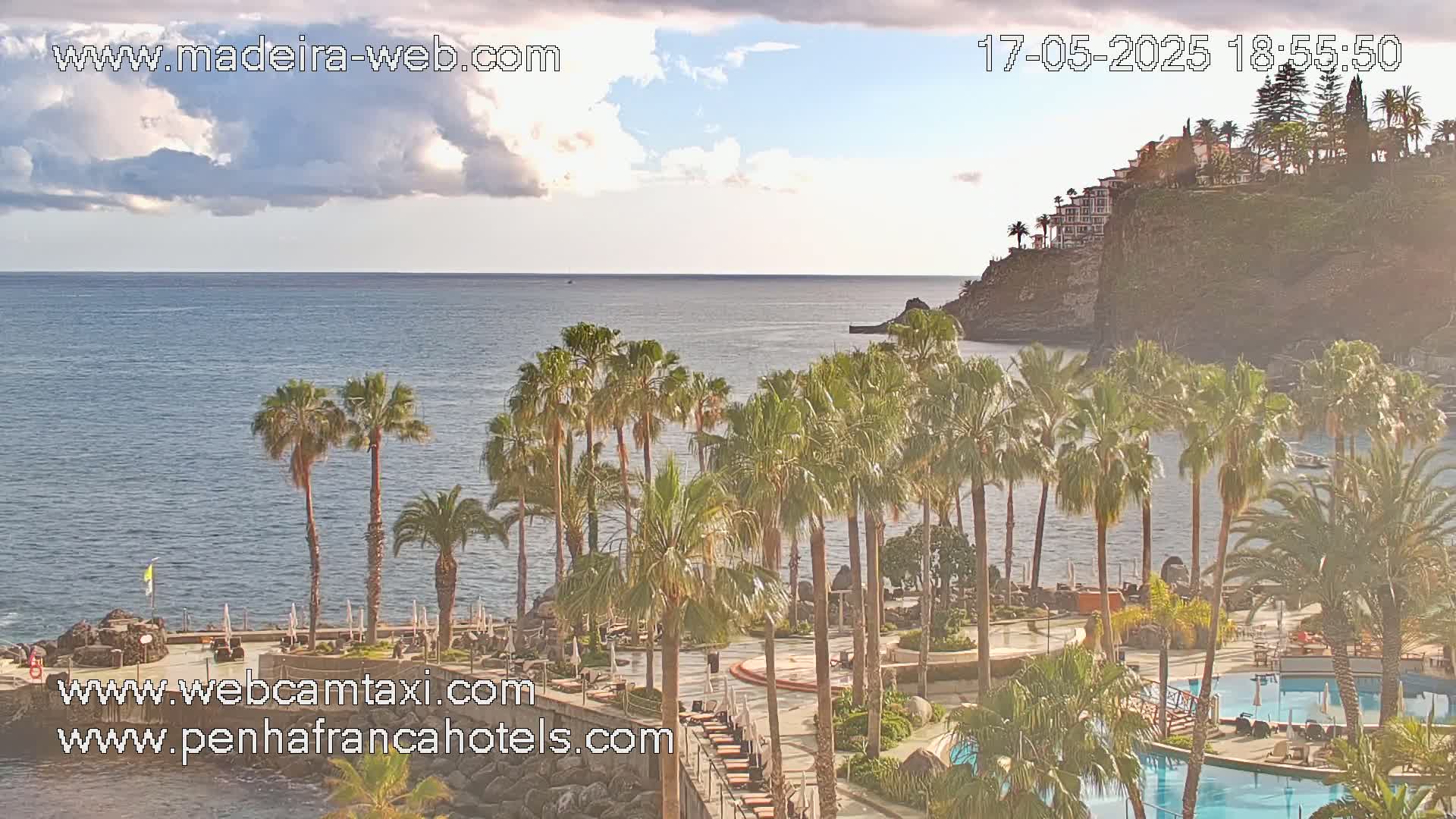 A partly cloudy day reveals a calm ocean, a clifftop hotel, and a resort area filled with palm trees and swimming pools.