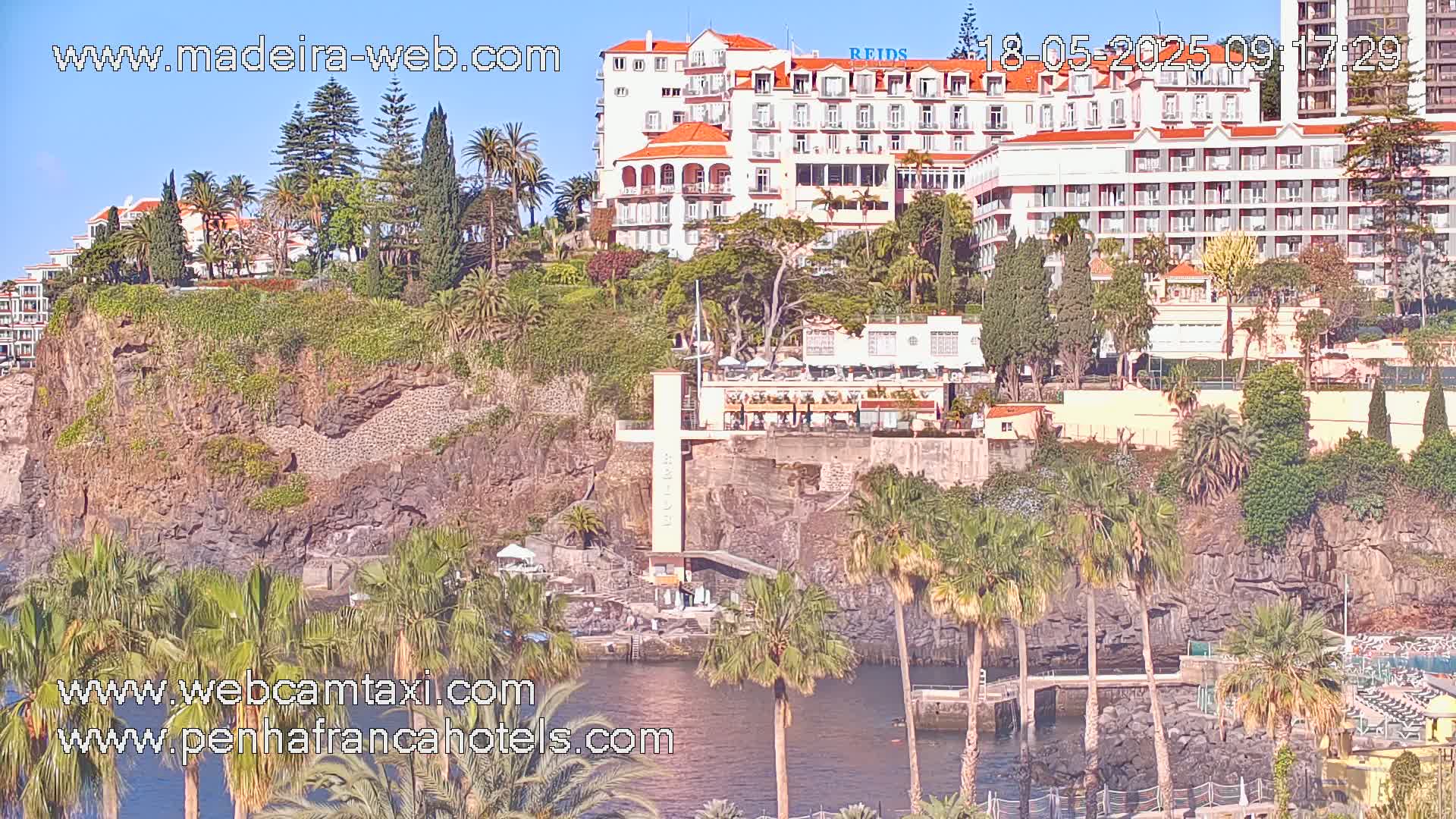 A coastal scene on a sunny day shows a large hotel complex built into a rocky cliffside, surrounded by palm trees and lush vegetation, with a calm body of water below.