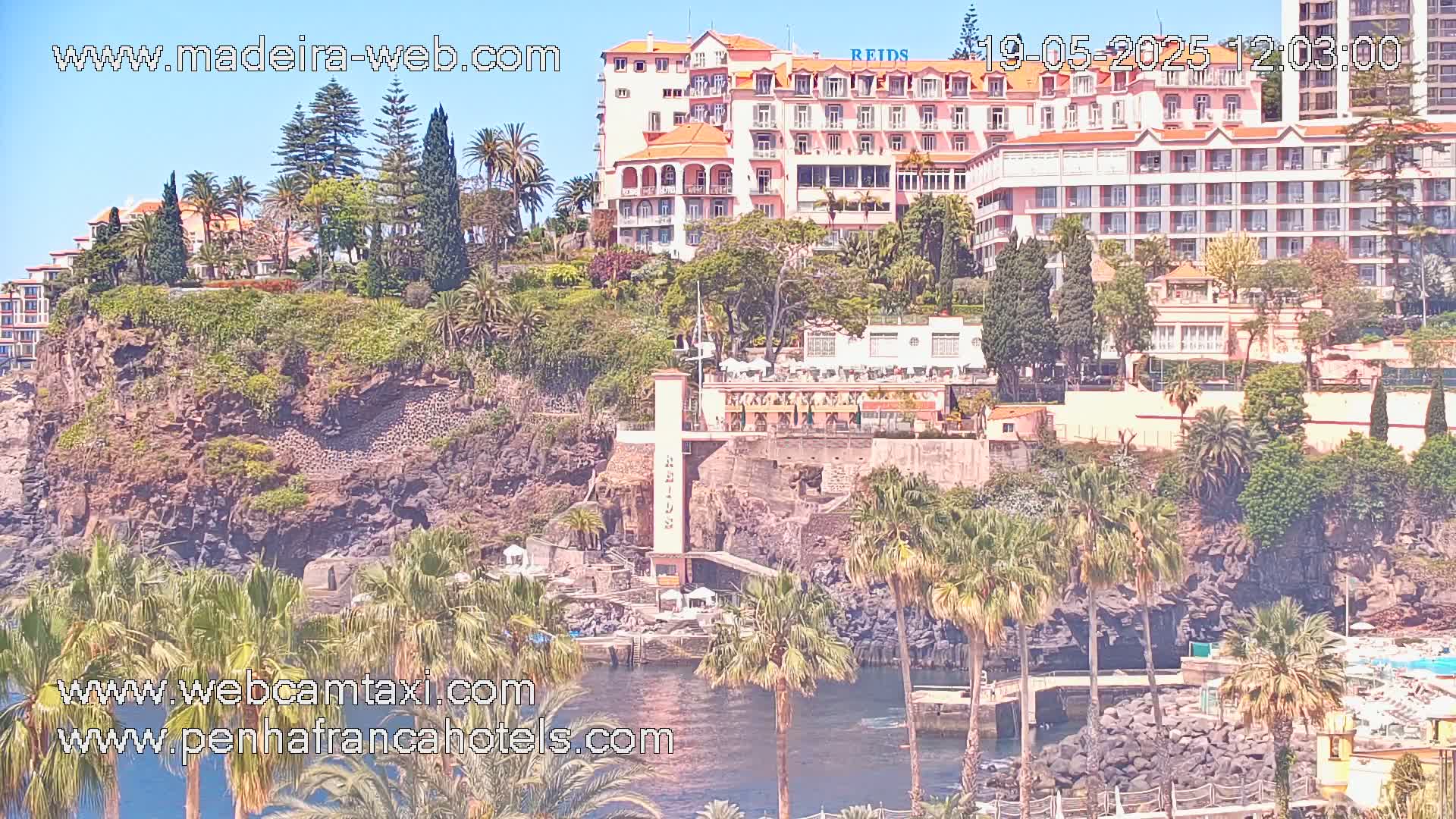 A large hotel complex sits atop a rocky cliff overlooking a calm body of water, surrounded by lush tropical vegetation under sunny skies.