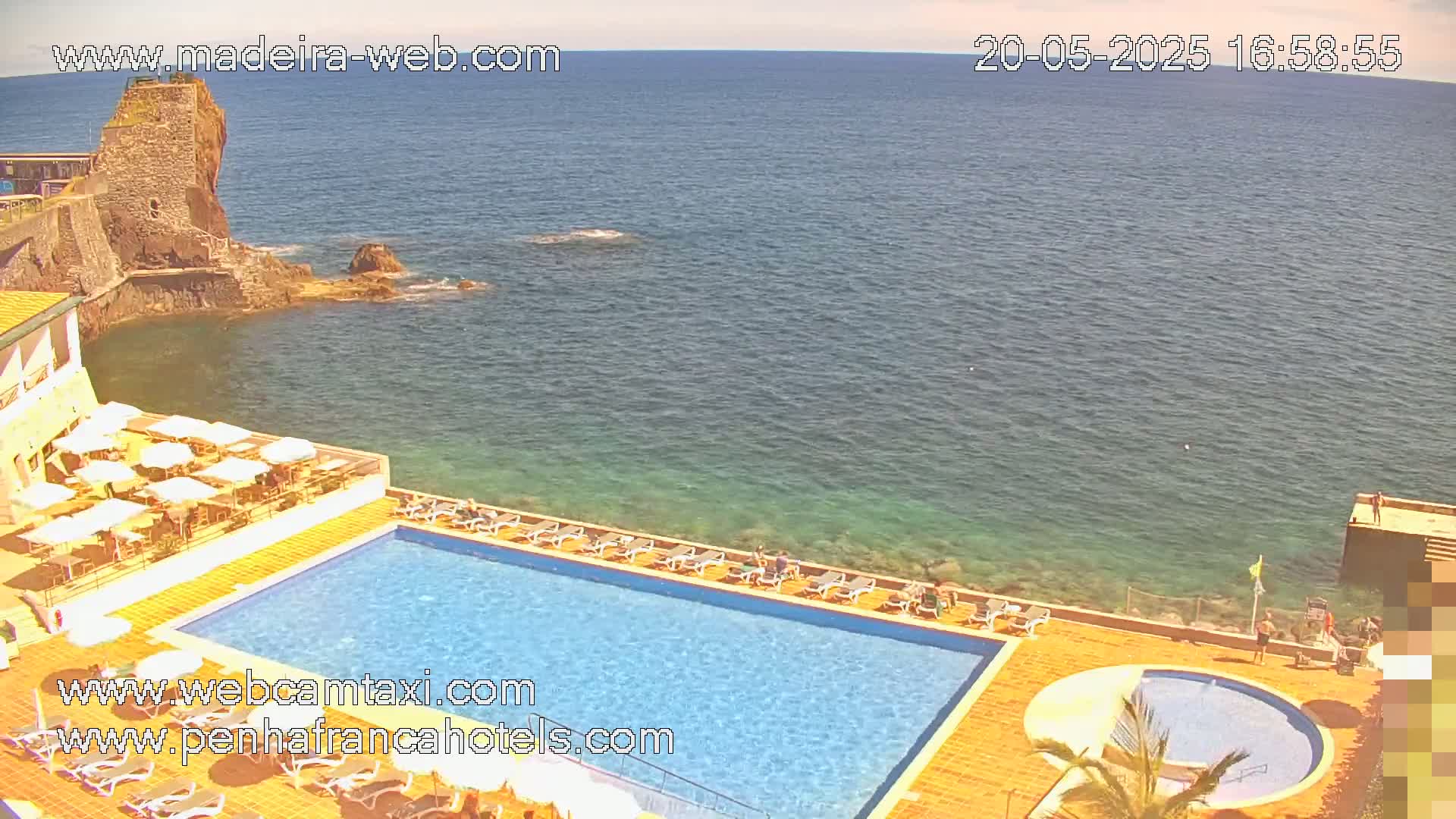 An outdoor pool and patio area overlook a calm ocean with a rocky coastline featuring a stone structure.
