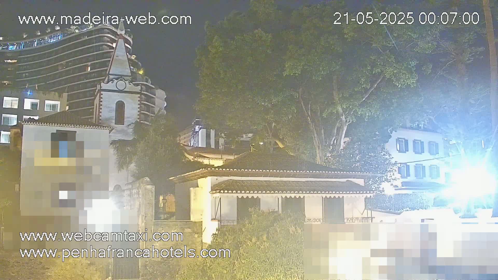 At night, a nighttime view shows several buildings, including a tall modern building, a smaller, older building with a steeple, and a low, single-story house, all surrounded by lush green trees.