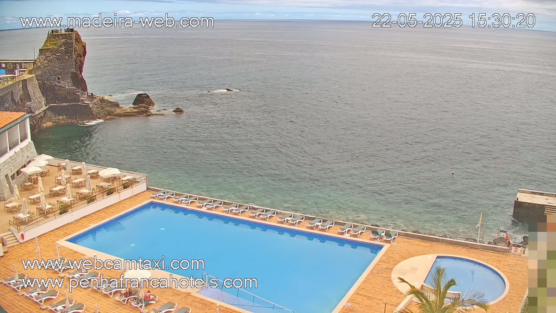 An outdoor swimming pool and patio area at a resort overlook a calm ocean and rocky coastline under a partly cloudy sky.